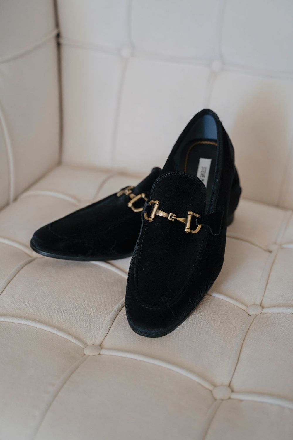 Black velvet loafers with gold hardware displayed on a tufted cream chair.