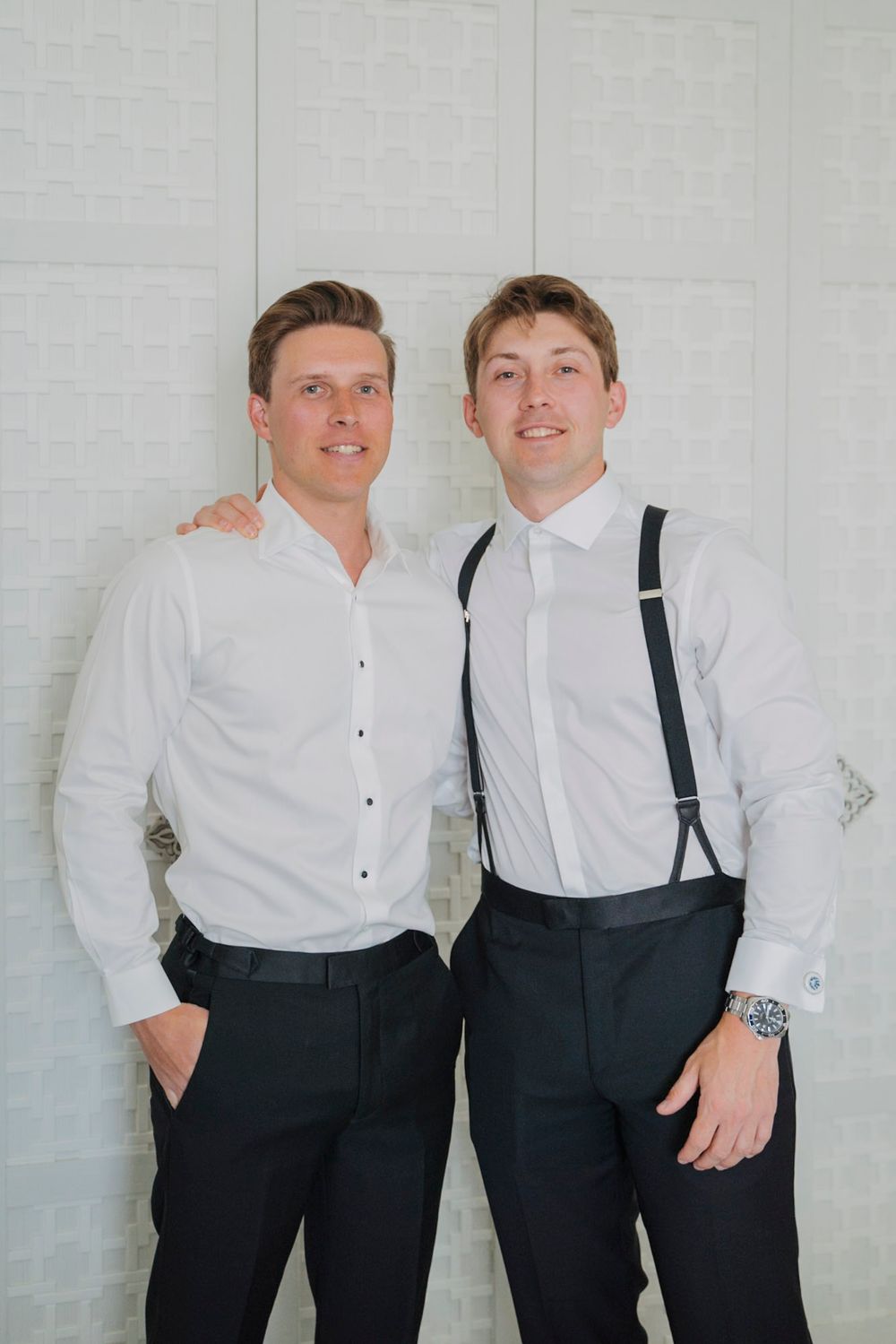 Two men in formal white shirts and dress pants posing against a white wall.