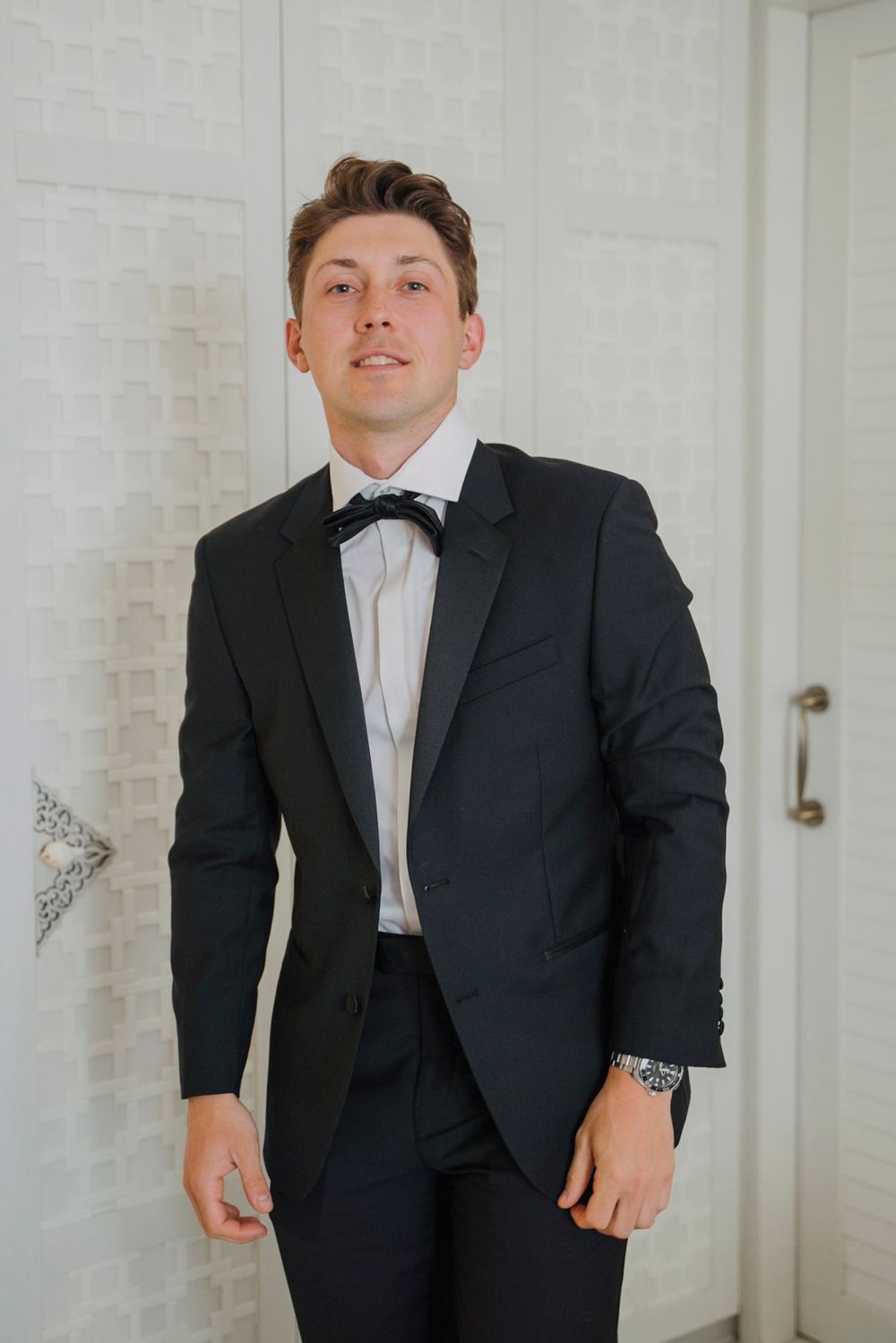 Group of groomsmen in black tuxedos posing against a white wall.
