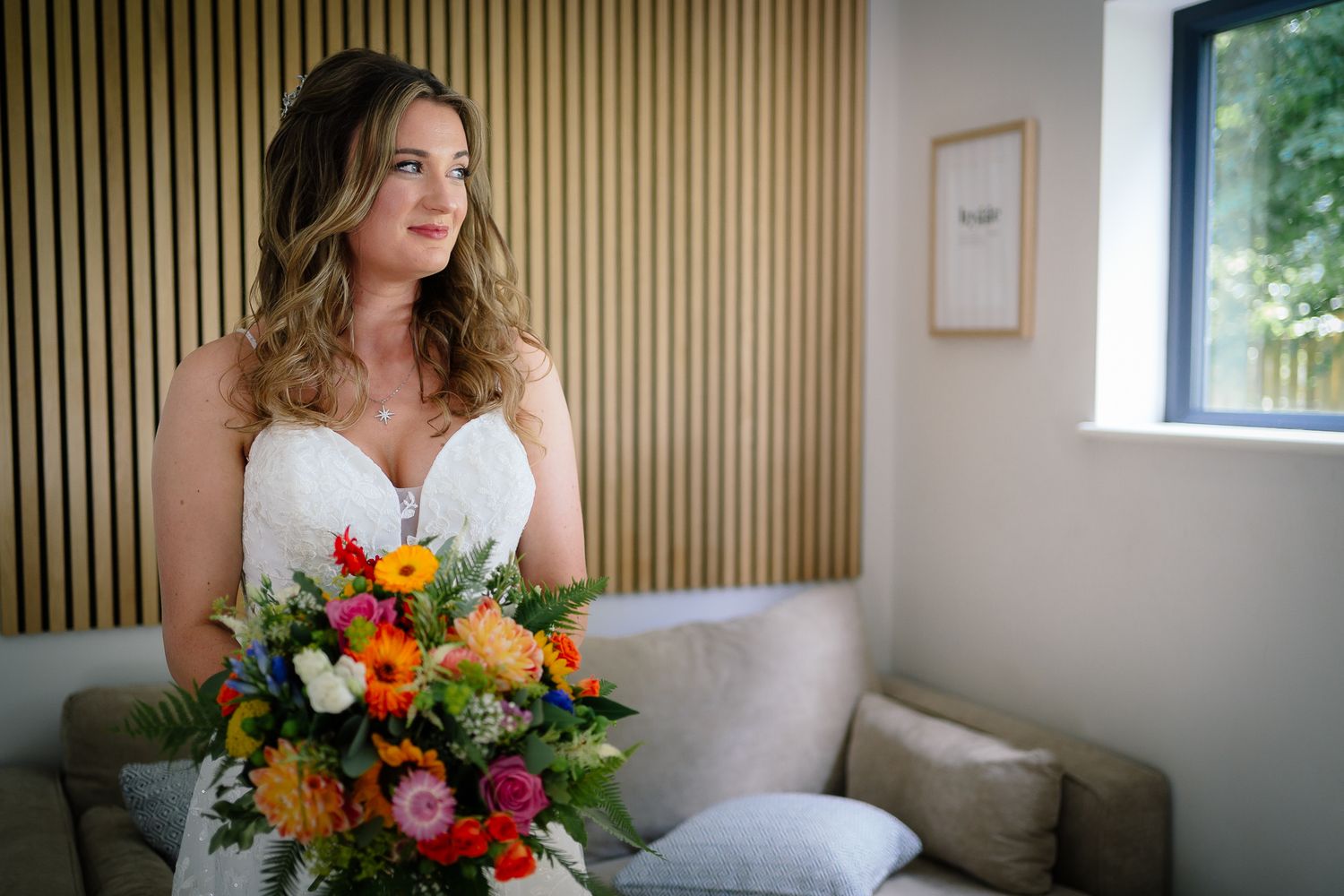 A bride in a white wedding dress holds a vibrant bouquet of orange, pink and purple flowers against a wood paneled wall.