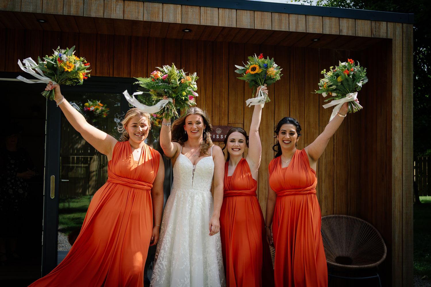 Wedding party poses together wearing coral orange bridesmaid dresses and white bridal gown against rustic barn backdrop.