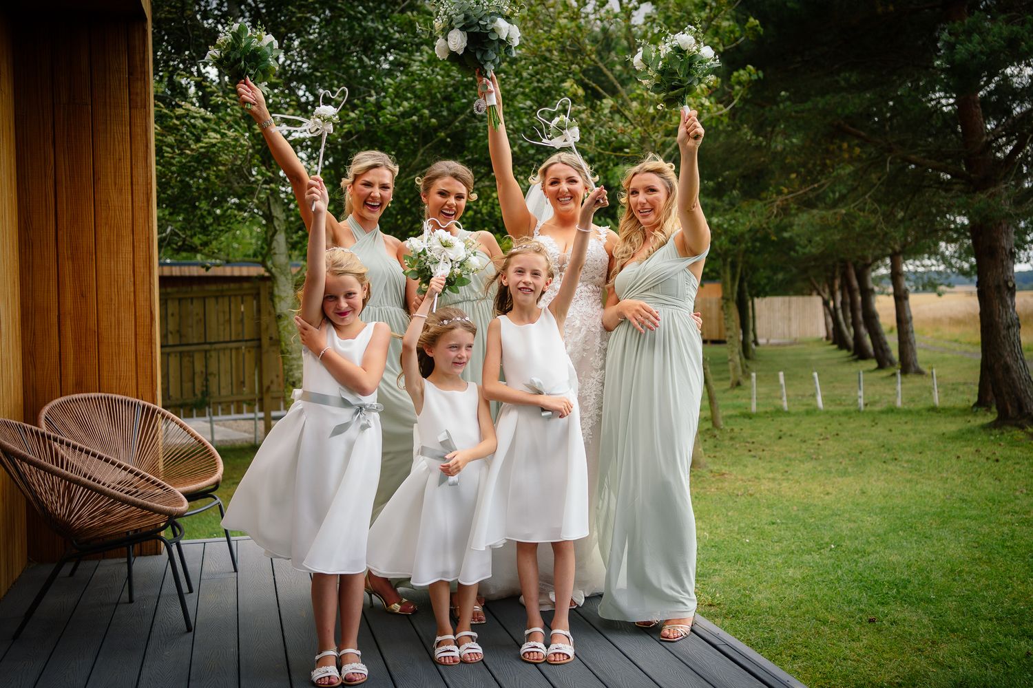 Group of bridesmaids in white dresses celebrating on a sunny lawn with raised arms.