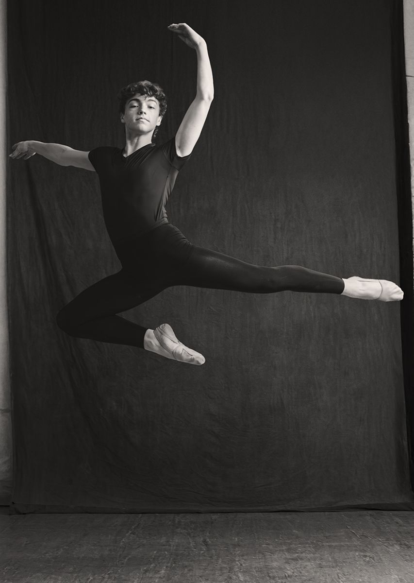 Male ballet dancer leaping gracefully in black attire and pointe shoes against dark backdrop in black and white photo.