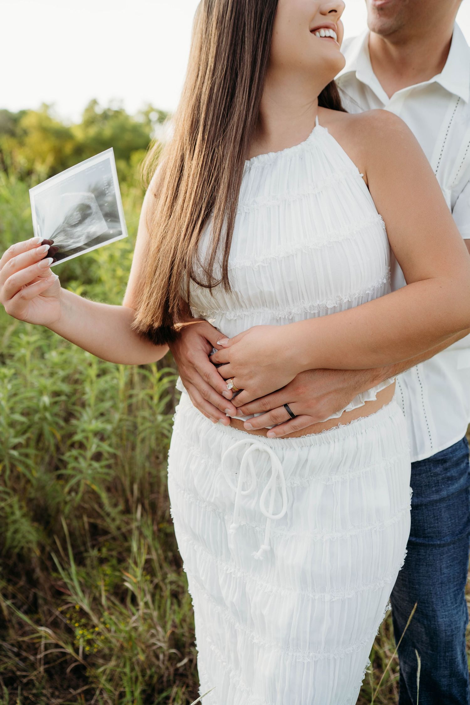 Couple in white dress and casual wear embrace while holding ultrasound photo outdoors in natural setting.