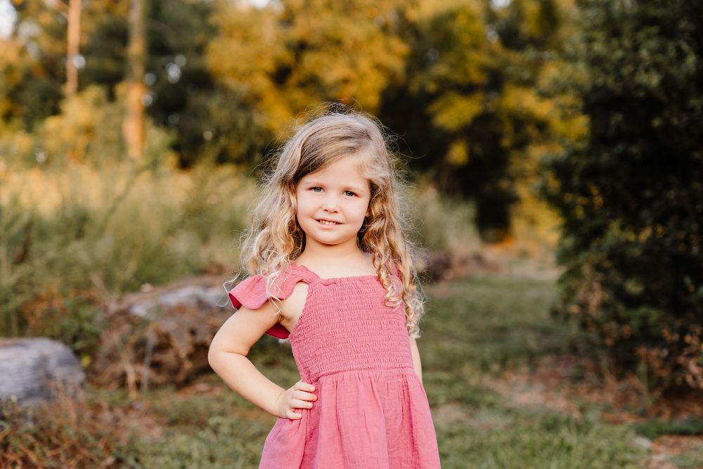 A child in a pink dress stands outdoors against a backdrop of autumn foliage and golden light.