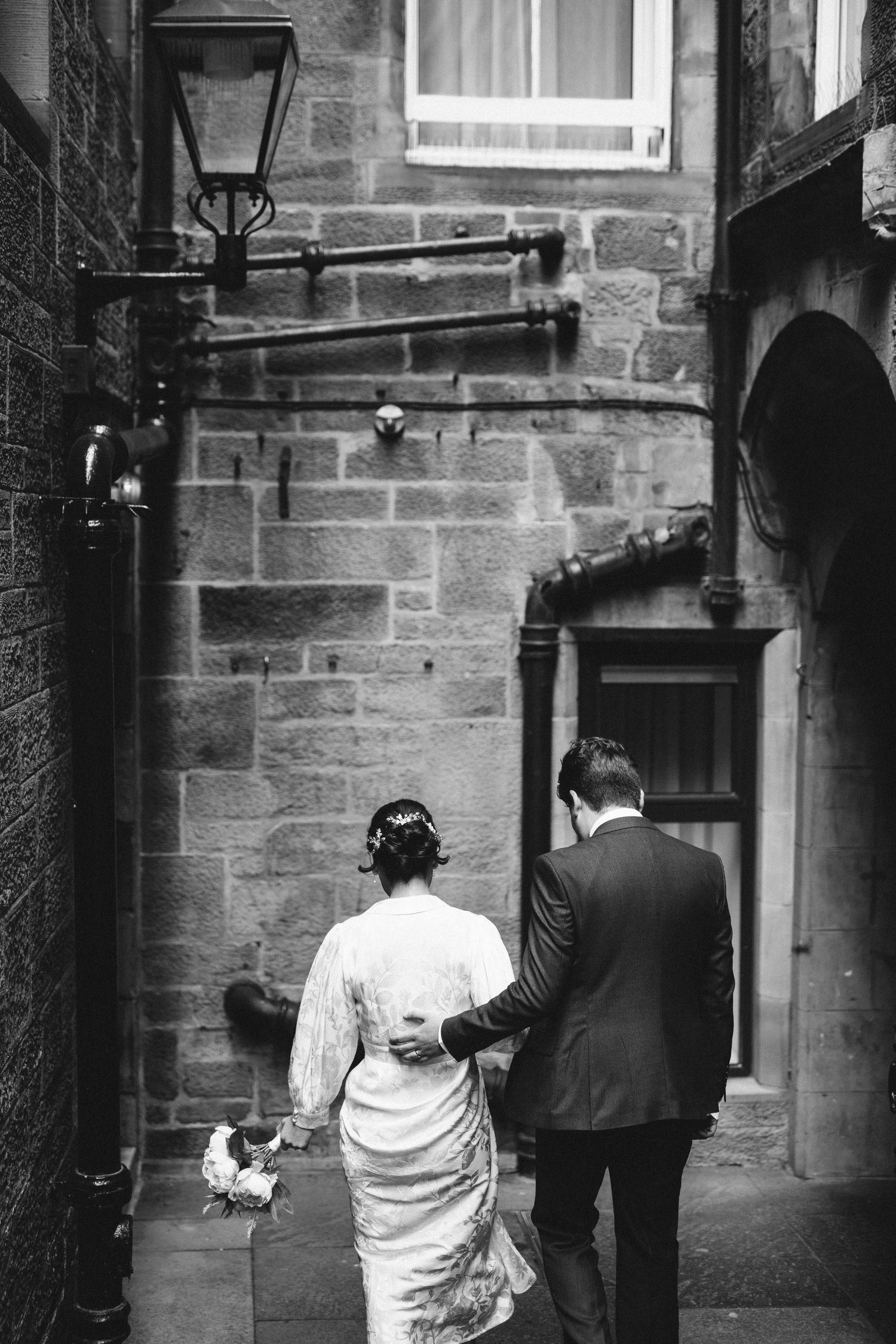 A couple walking down a narrow stone alleyway with vintage street lamps in black and white.