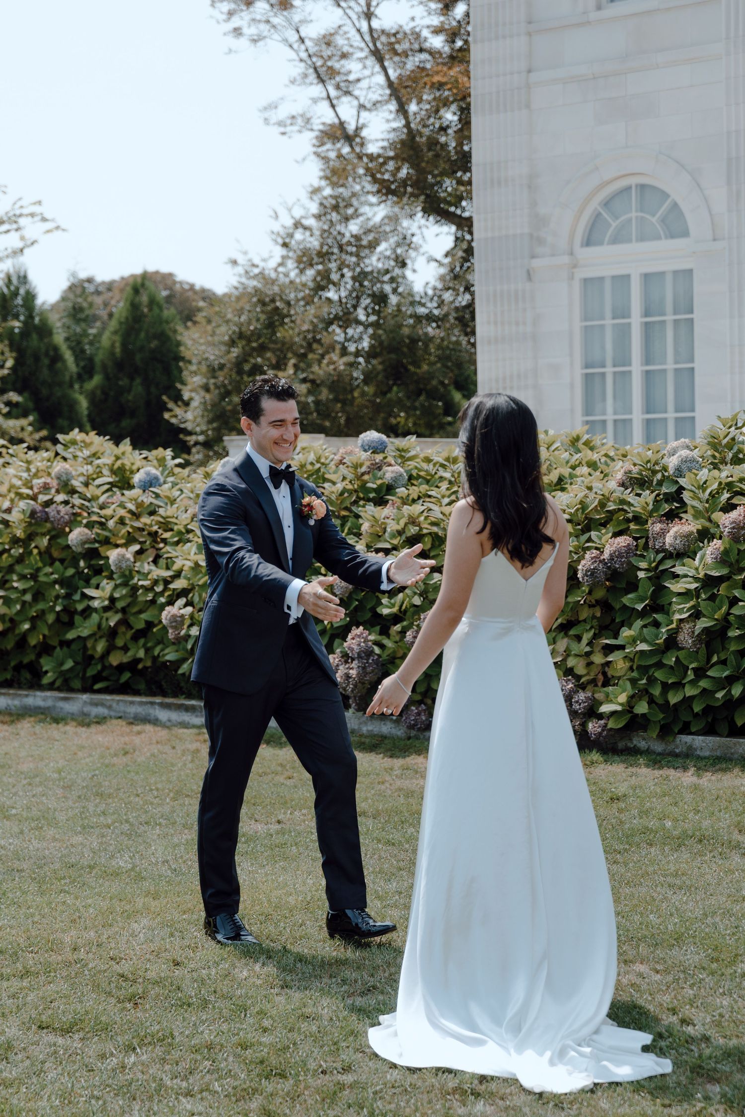 A couple in wedding attire shares a playful moment together in a garden setting with white architectural details.