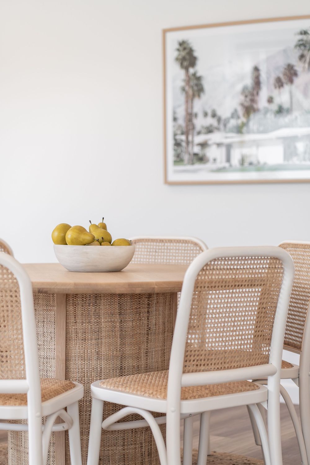 Modern coastal dining room with rattan chairs, light wood table, bowl of pears, and framed palm tree artwork on wall.