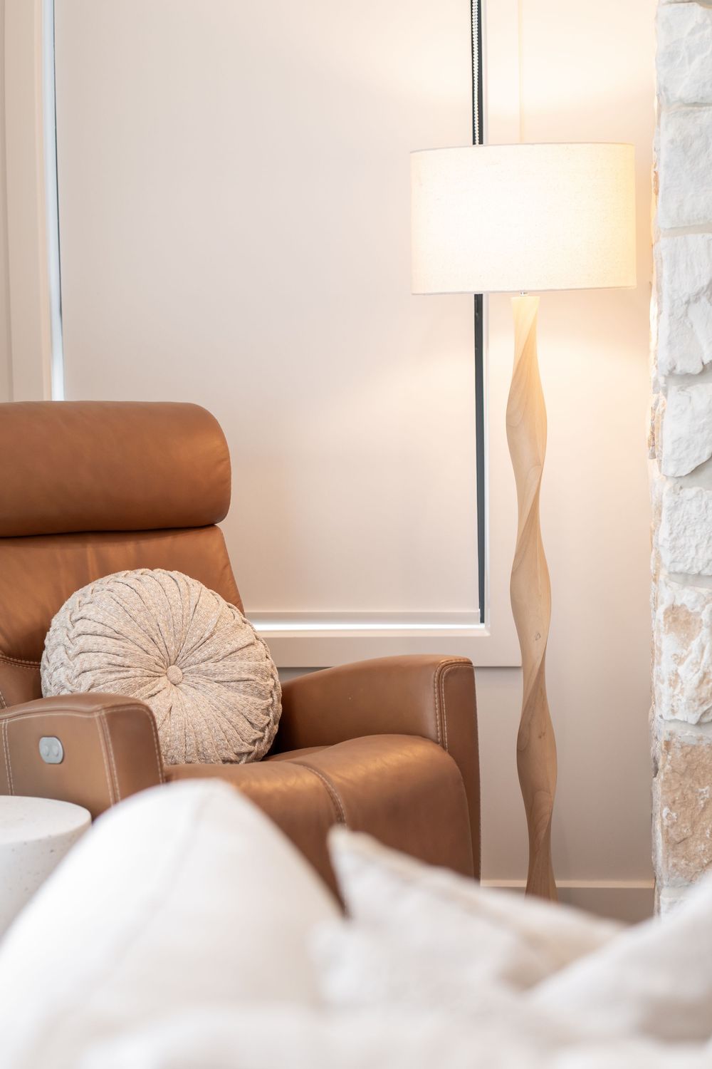 Cozy brown leather armchair with round beige pillow next to window with white curtains and table lamp.