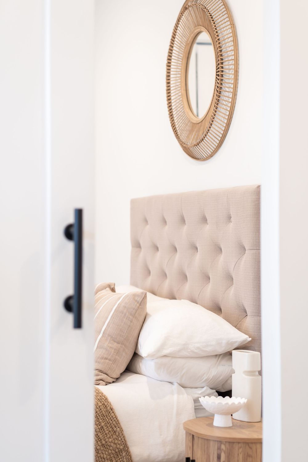 Elegant bedroom interior with beige tufted headboard, white bedding, and circular rattan wall decor above.