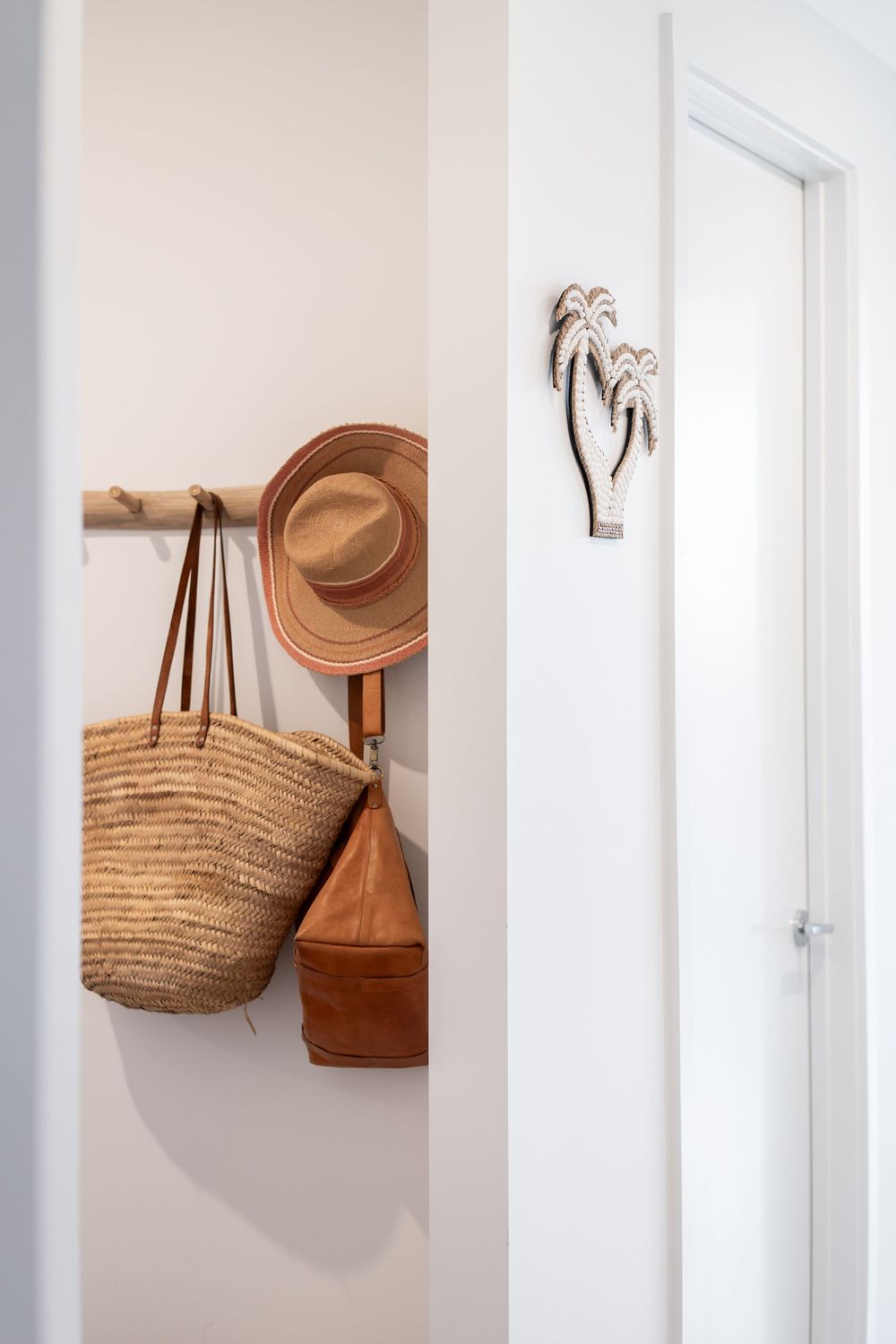 A straw beach bag and brown leather purse hang on minimalist white wall hooks against a bright white wall.
