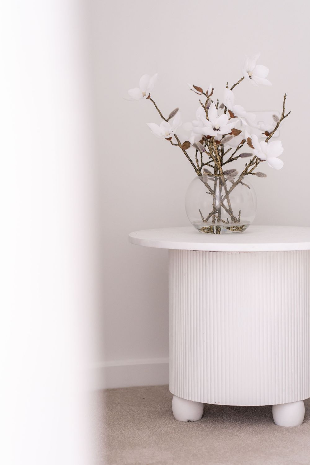 White ceramic vase with delicate white flowers sits atop a textured cylindrical pedestal against a light background.