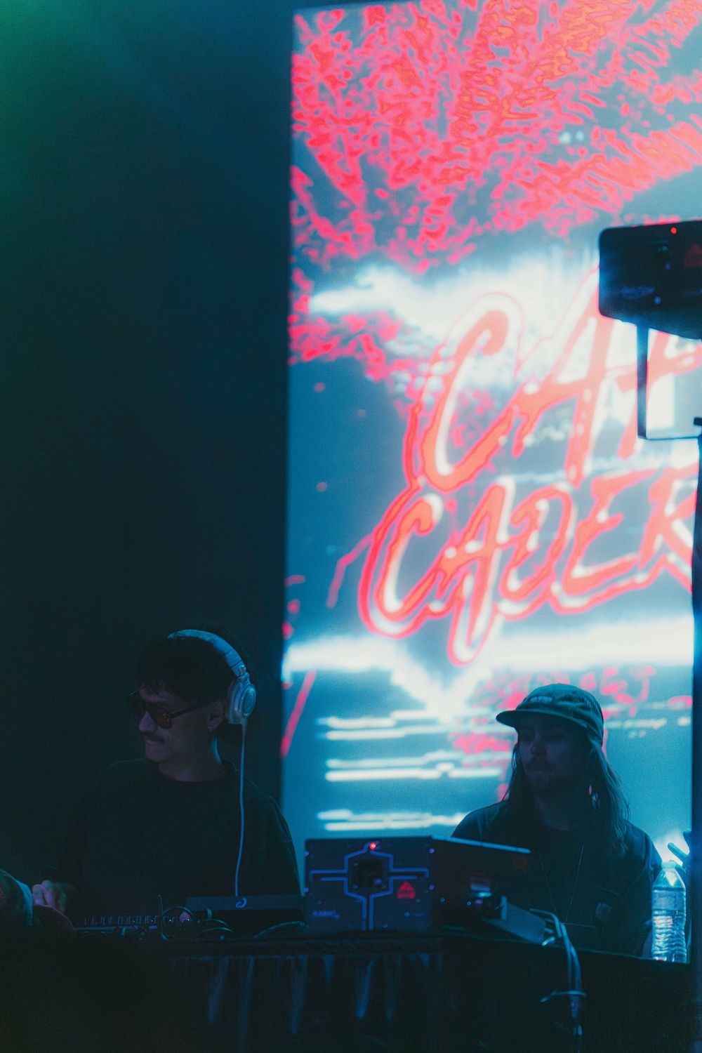 DJs perform at a nightclub with neon pink 'Cafe' signage glowing against a blue backdrop in atmospheric lighting.