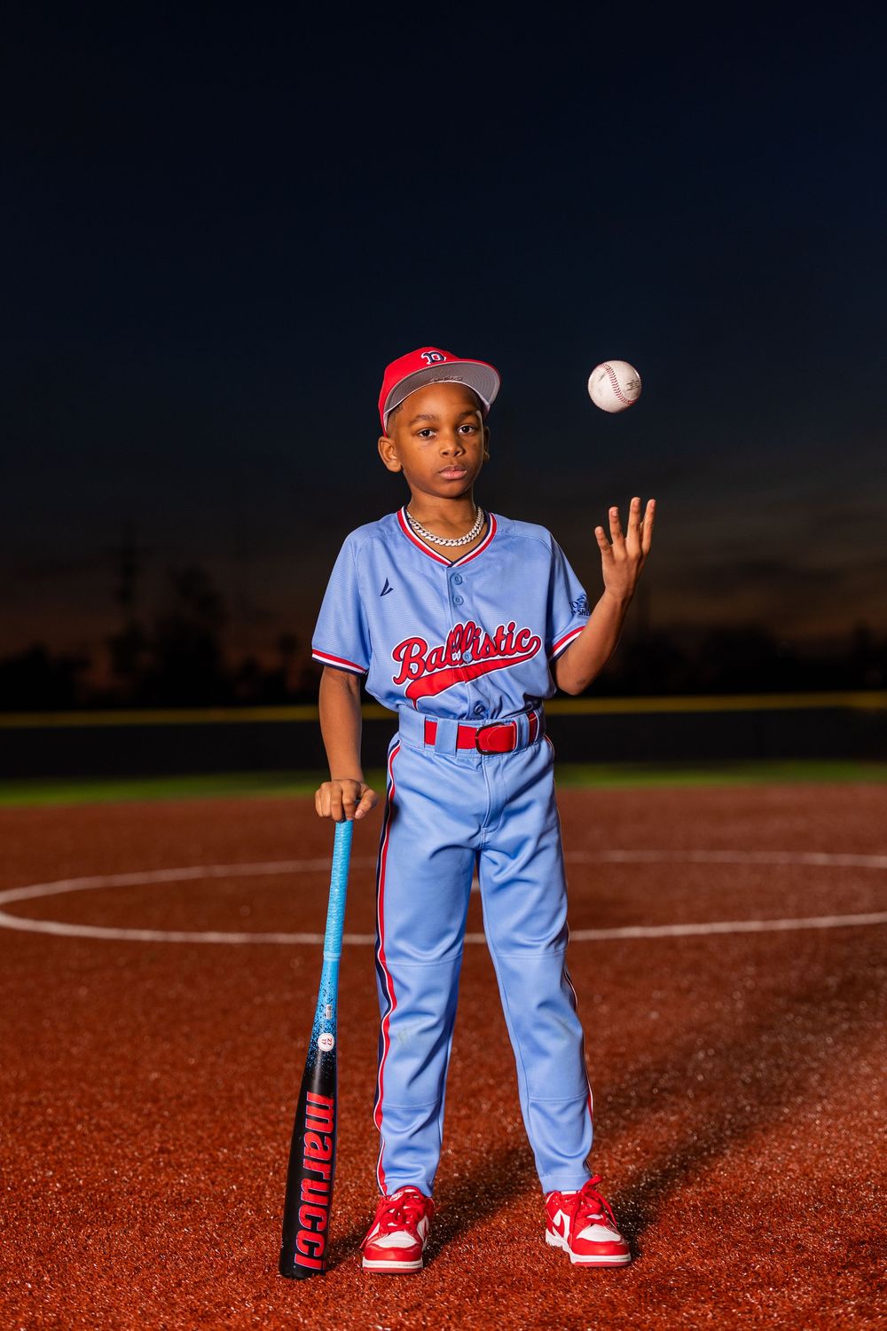 Young baseball player in light blue uniform poses with ball and bat on dirt field at dusk.