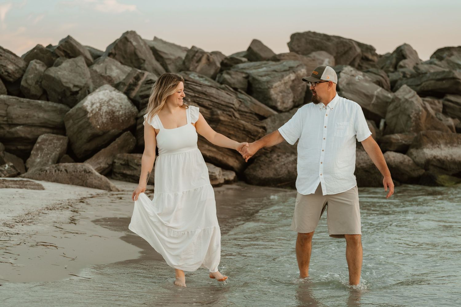A couple holds hands while walking along a rocky beach at sunset wearing white and casual summer attire.