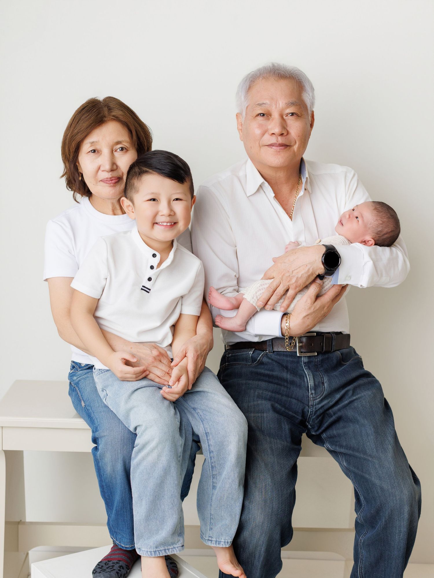 A family wearing white shirts and jeans sits together on a white bench in a minimalist studio setting.