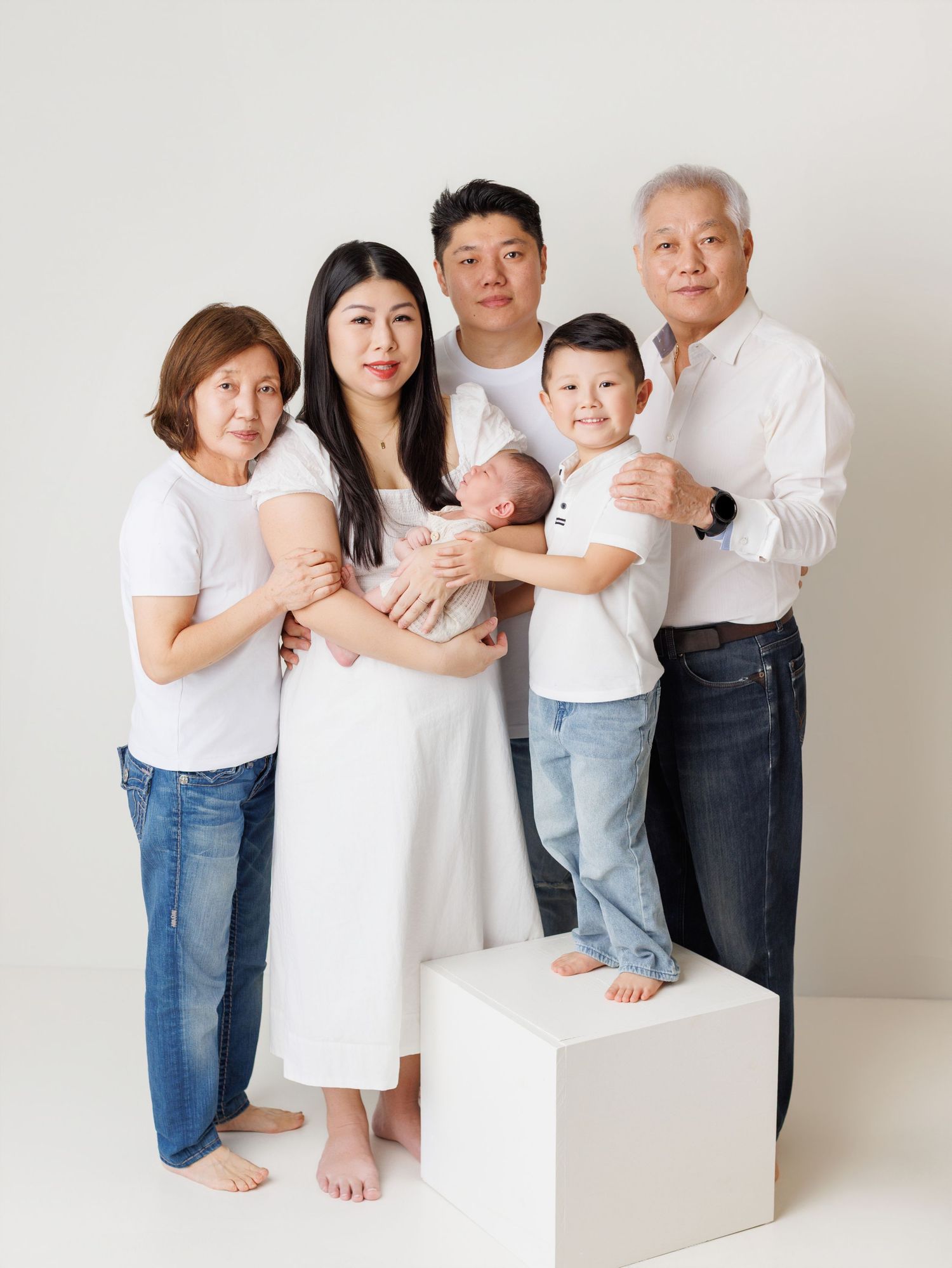 A multi-generational family wearing white tops and jeans poses together on white blocks in a minimalist studio setting.