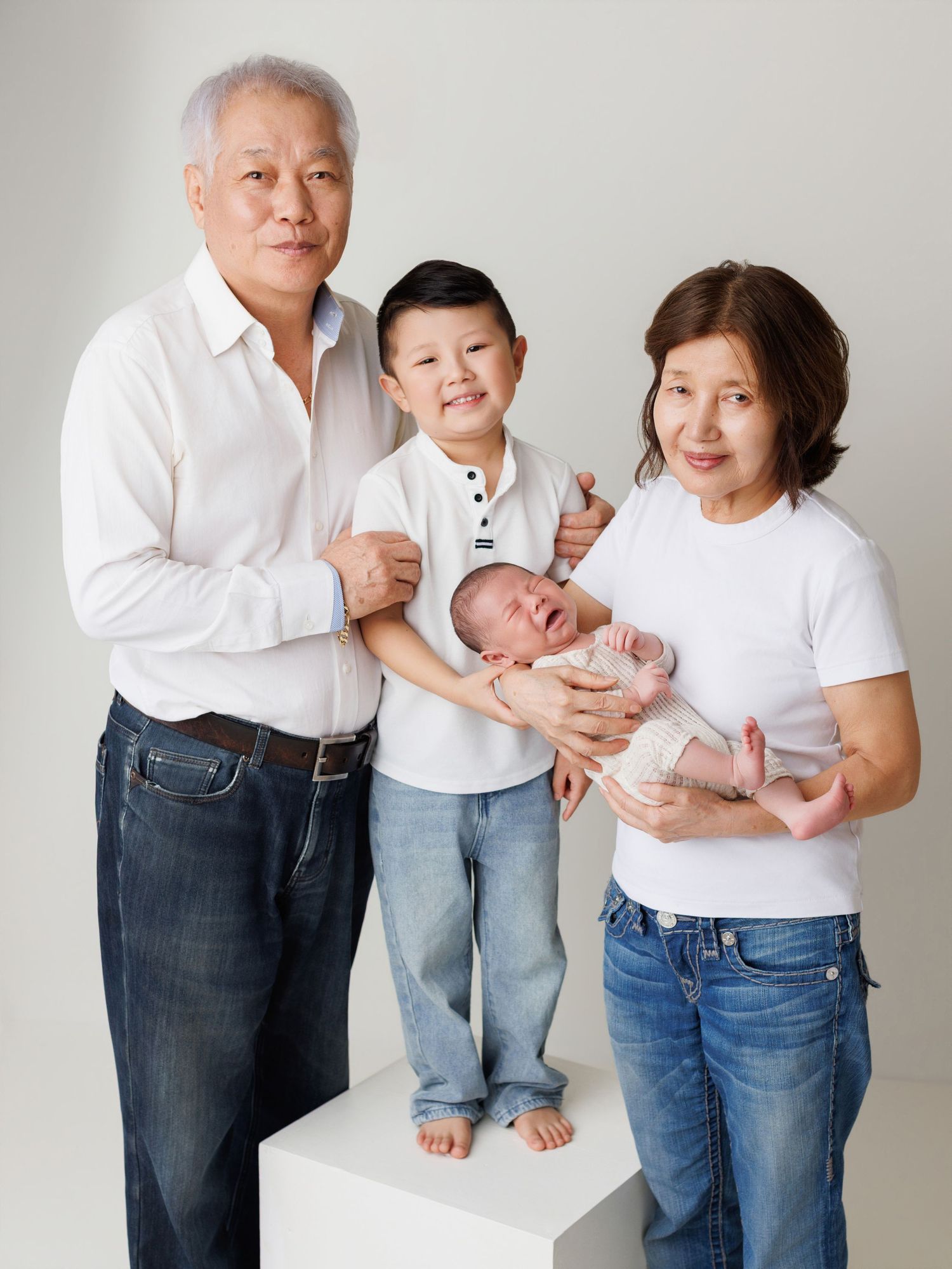 A family portrait against a white backdrop with a baby, young boy, and two adults in casual attire.