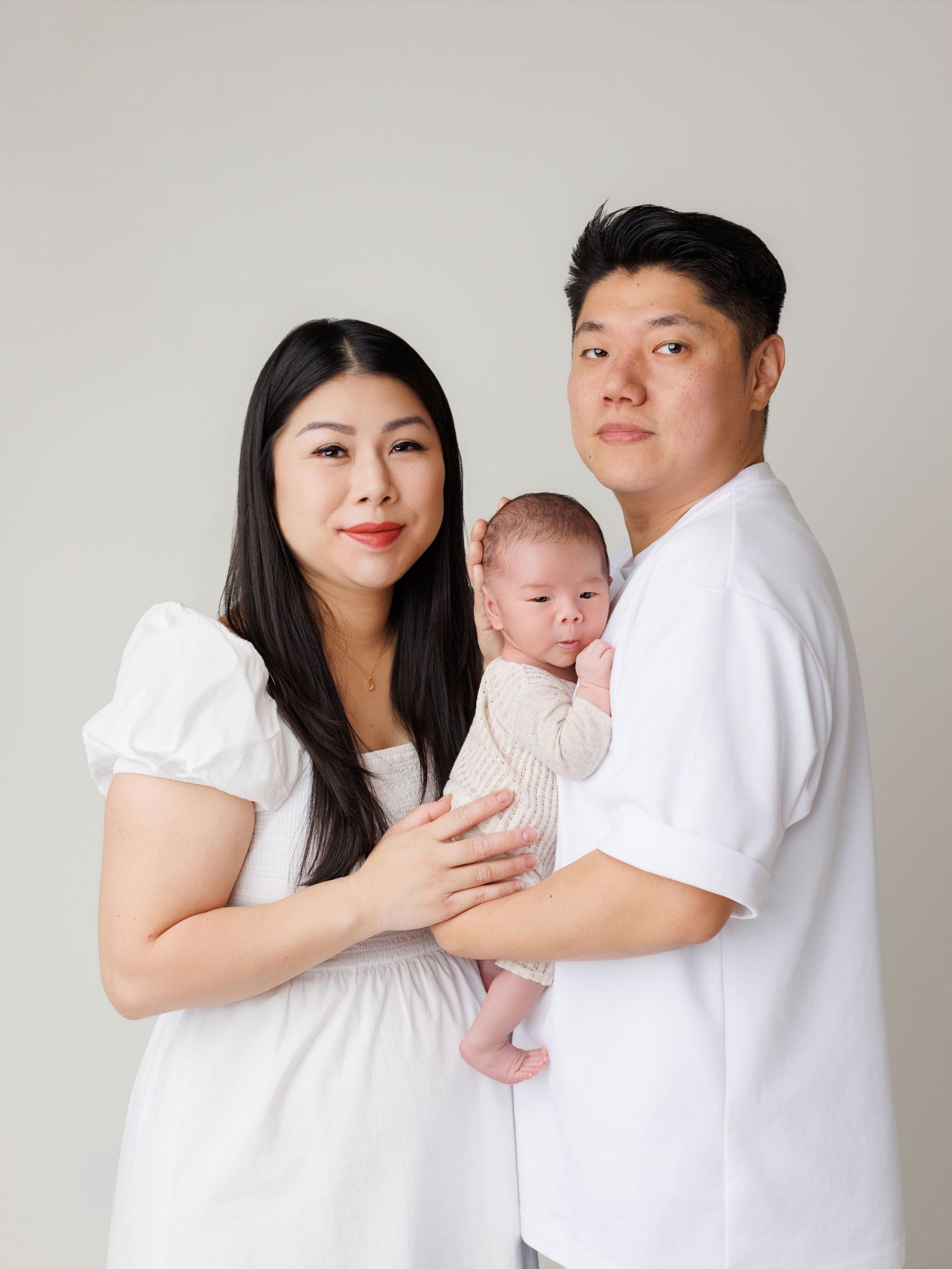 Family wearing white outfits poses together with newborn baby in minimalist studio setting.