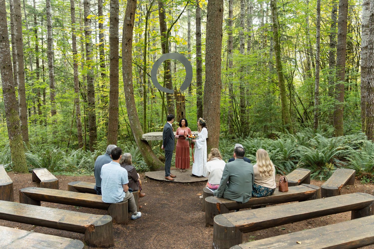 Visitors relax on wooden benches along a peaceful forest trail surrounded by tall trees and lush greenery.