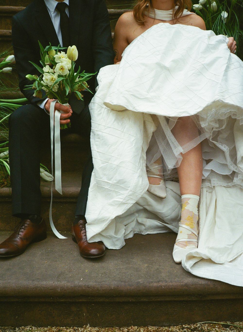 A close-up view of a bride's white wedding dress and groom's brown shoes as they sit together on stone steps.