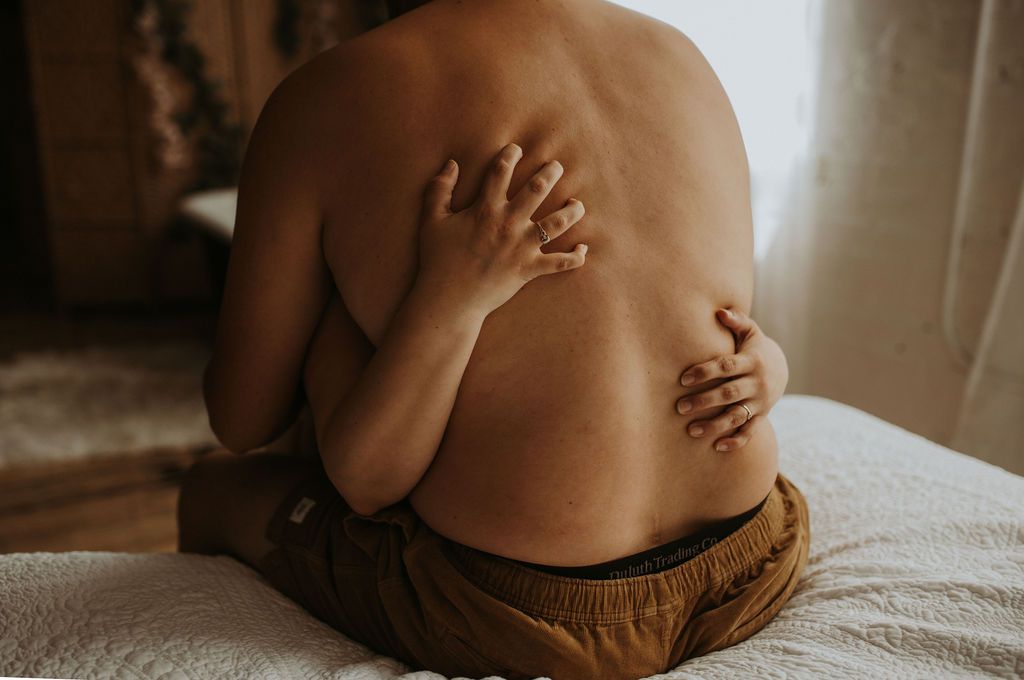 Young child sitting on a white bed, viewed from behind, wearing brown shorts with hands resting on their back.