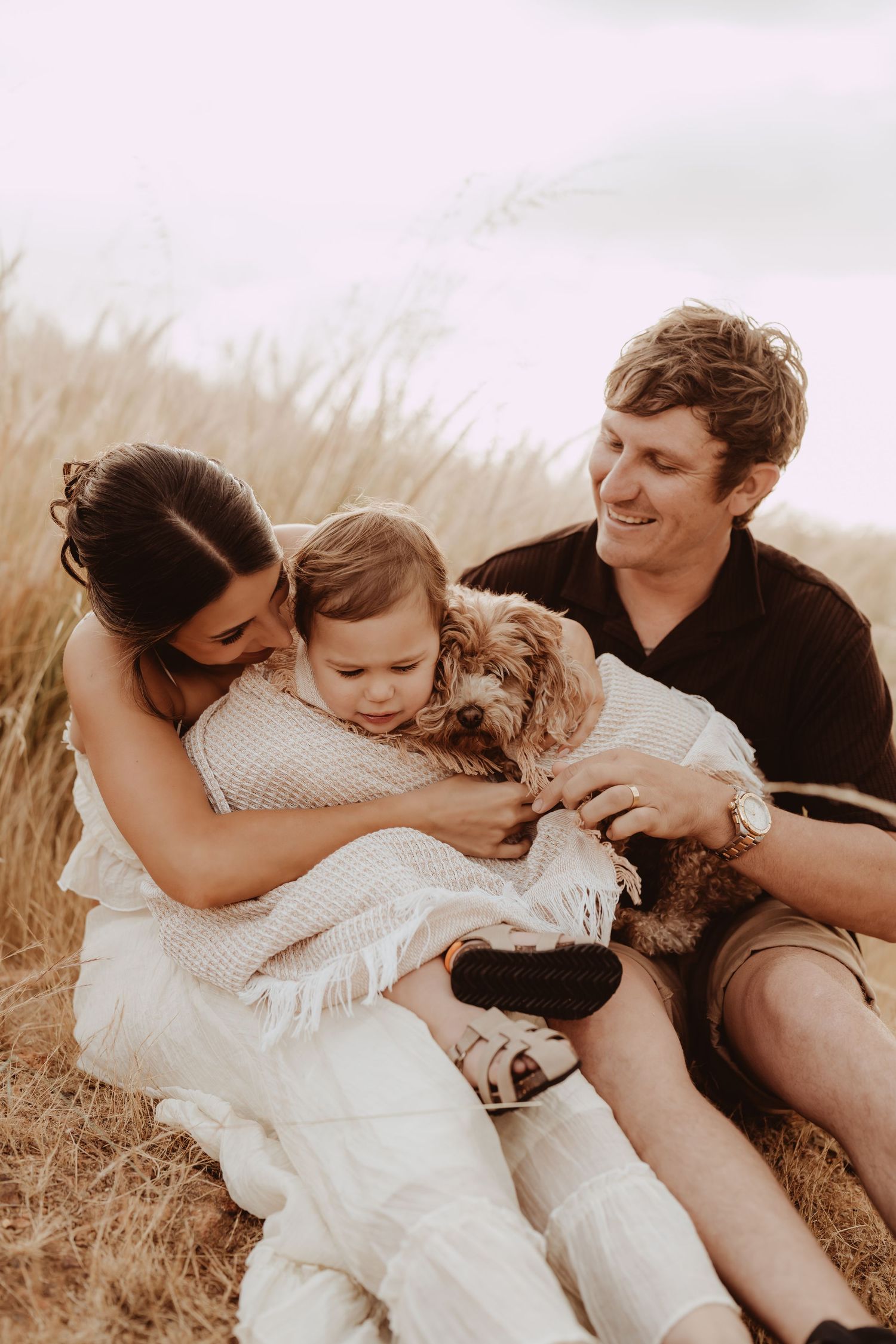 A pregnant woman in a white dress holds an ultrasound photo in a golden sunlit field at sunset.