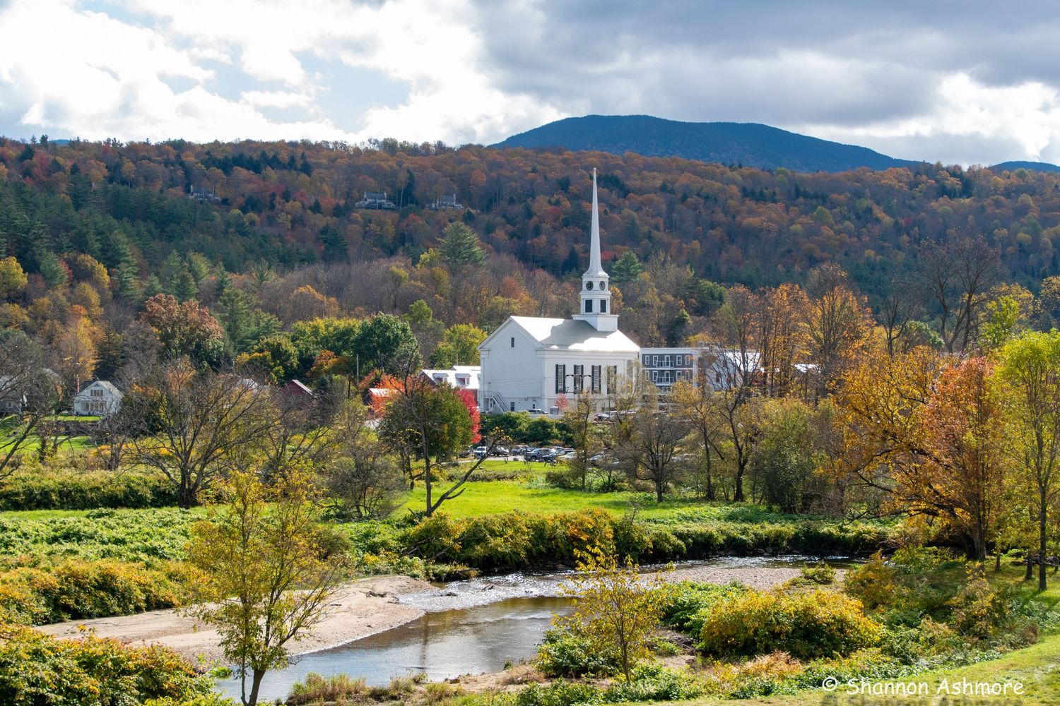 White steeple church nestled in Vermont village with river, fall foliage, and mountain backdrop.