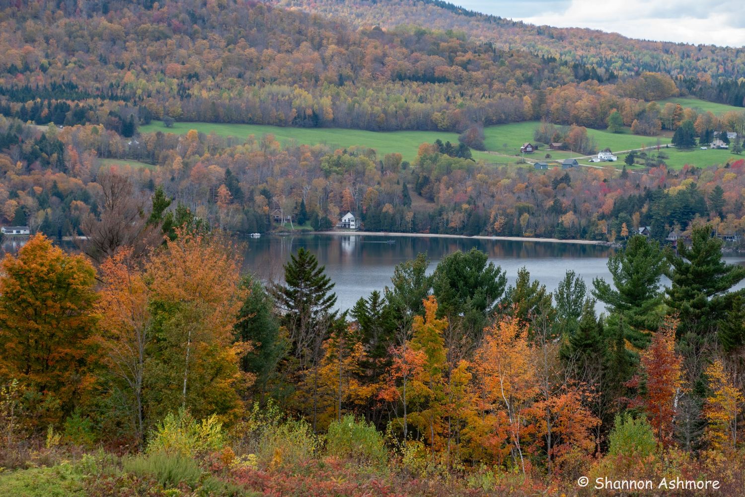 Aerial view of a serene lake surrounded by vibrant autumn foliage and rolling hills in New England.