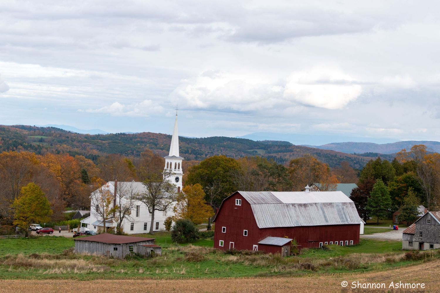 Classic red barn and white church surrounded by autumn foliage in scenic Vermont countryside.