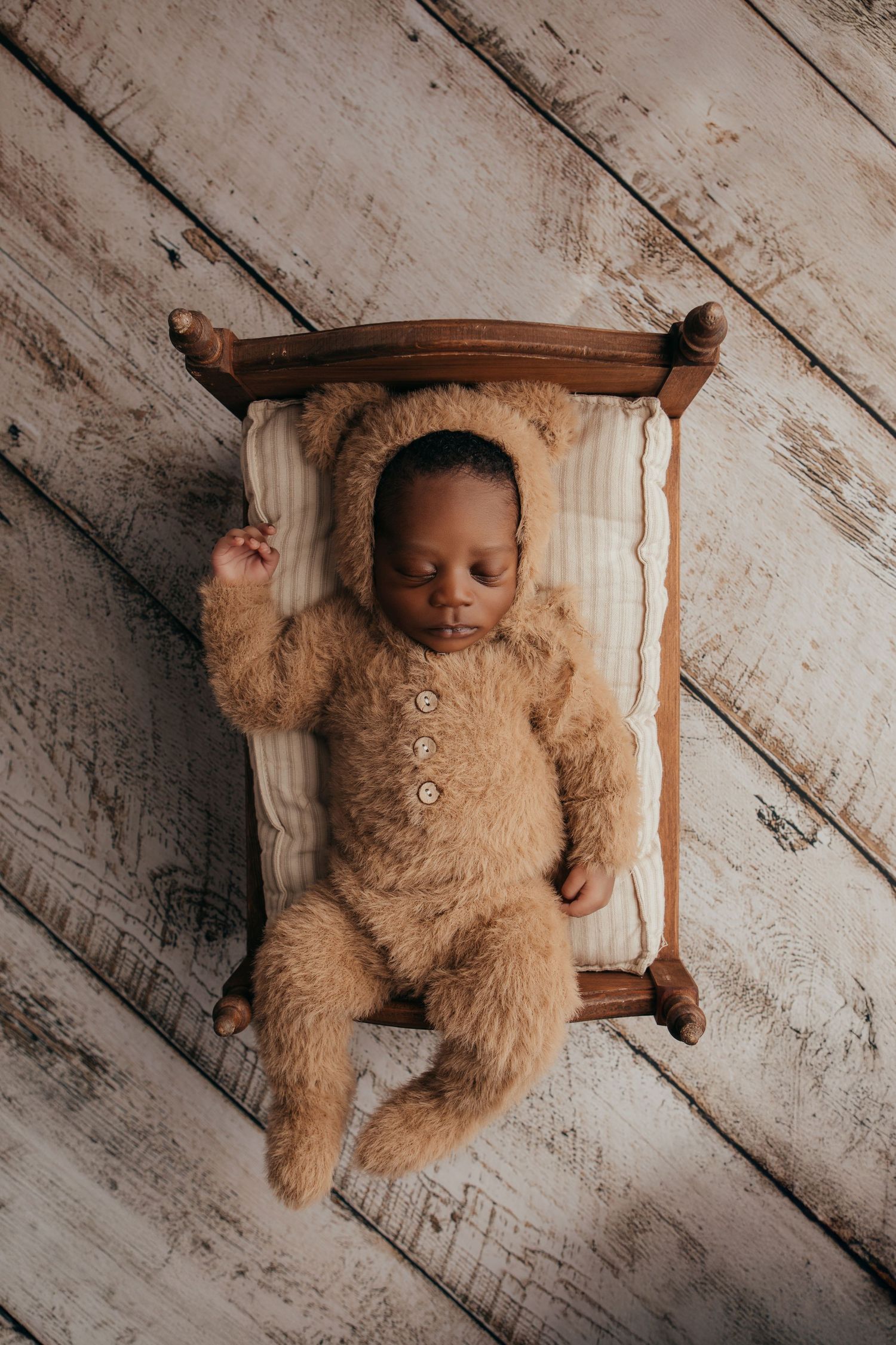 Baby wearing fuzzy brown teddy bear outfit lying on rustic wooden floor.