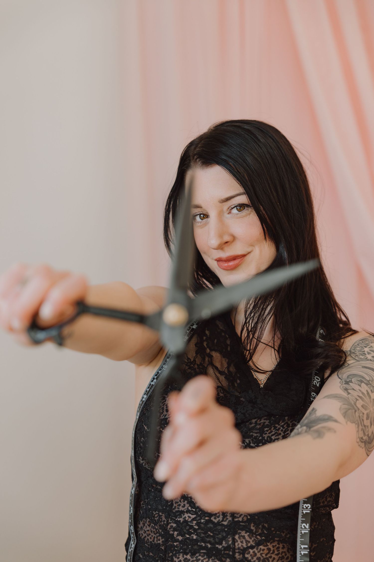 Dark-haired woman with tattoos holding large scissors toward camera wearing black lace top with measuring tape.