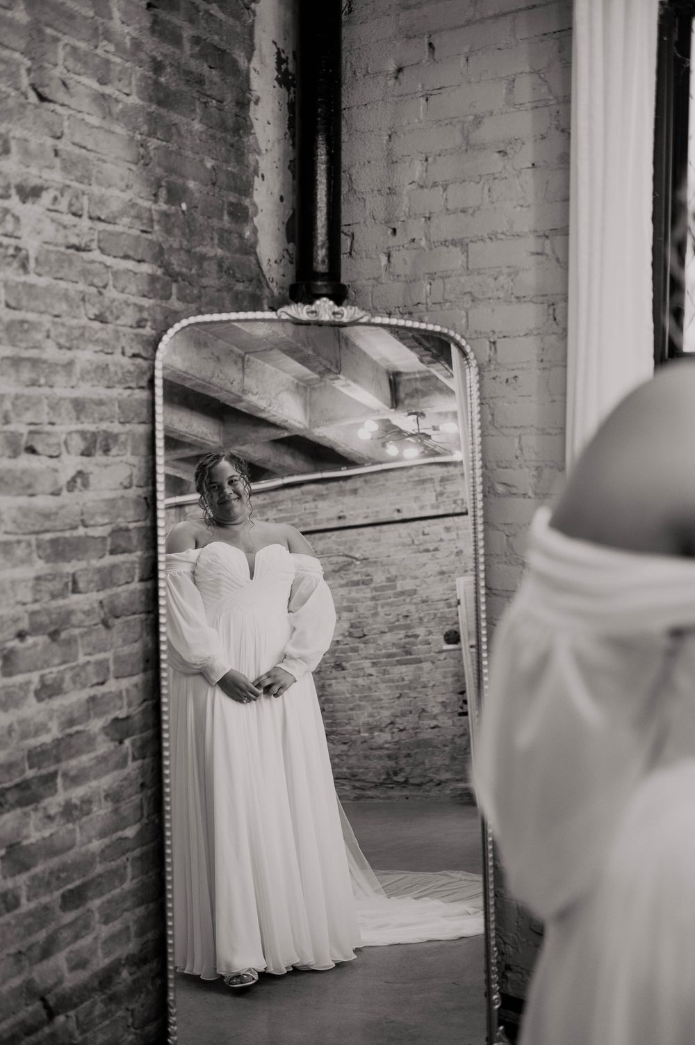 Black and white photo of bride in flowing white gown with puff sleeves admiring her reflection in an ornate floor mirror.