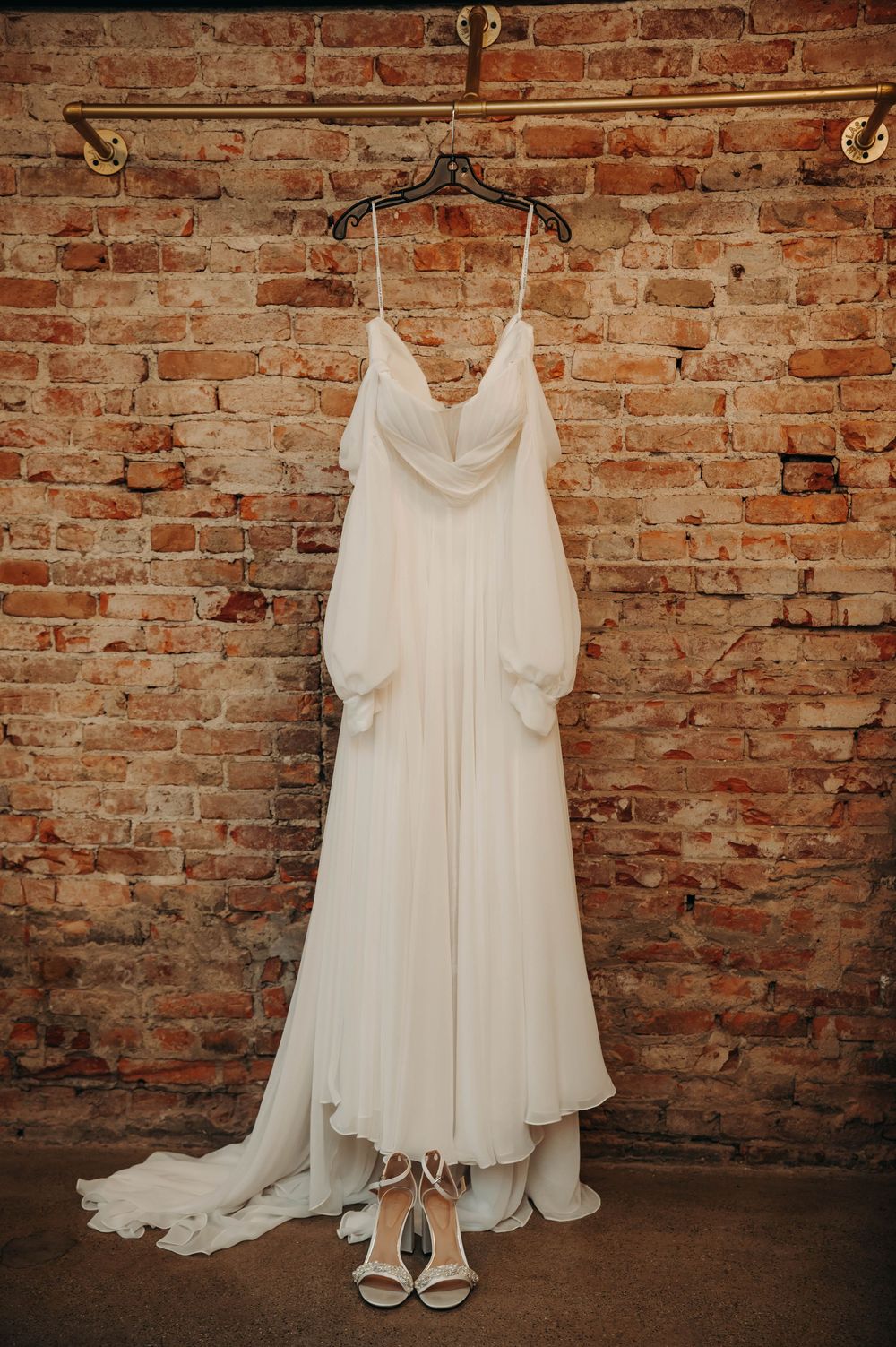 Elegant white wedding gown with puff sleeves hanging on a brass rack against a rustic exposed brick wall with bridal heels below.