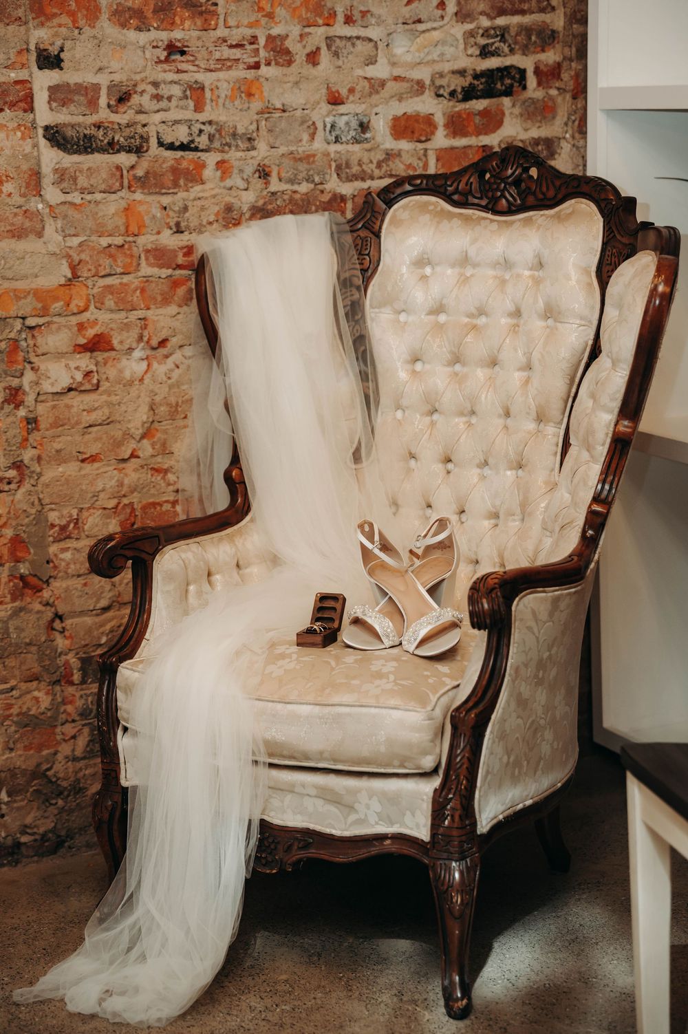 Vintage tufted wingback chair with bridal veil and white strappy wedding heels against exposed brick wall.