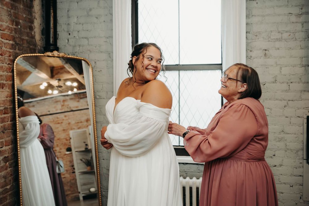 Smiling bride in off-shoulder white gown laughs with a woman in pink dress helping her get ready near a mirror.