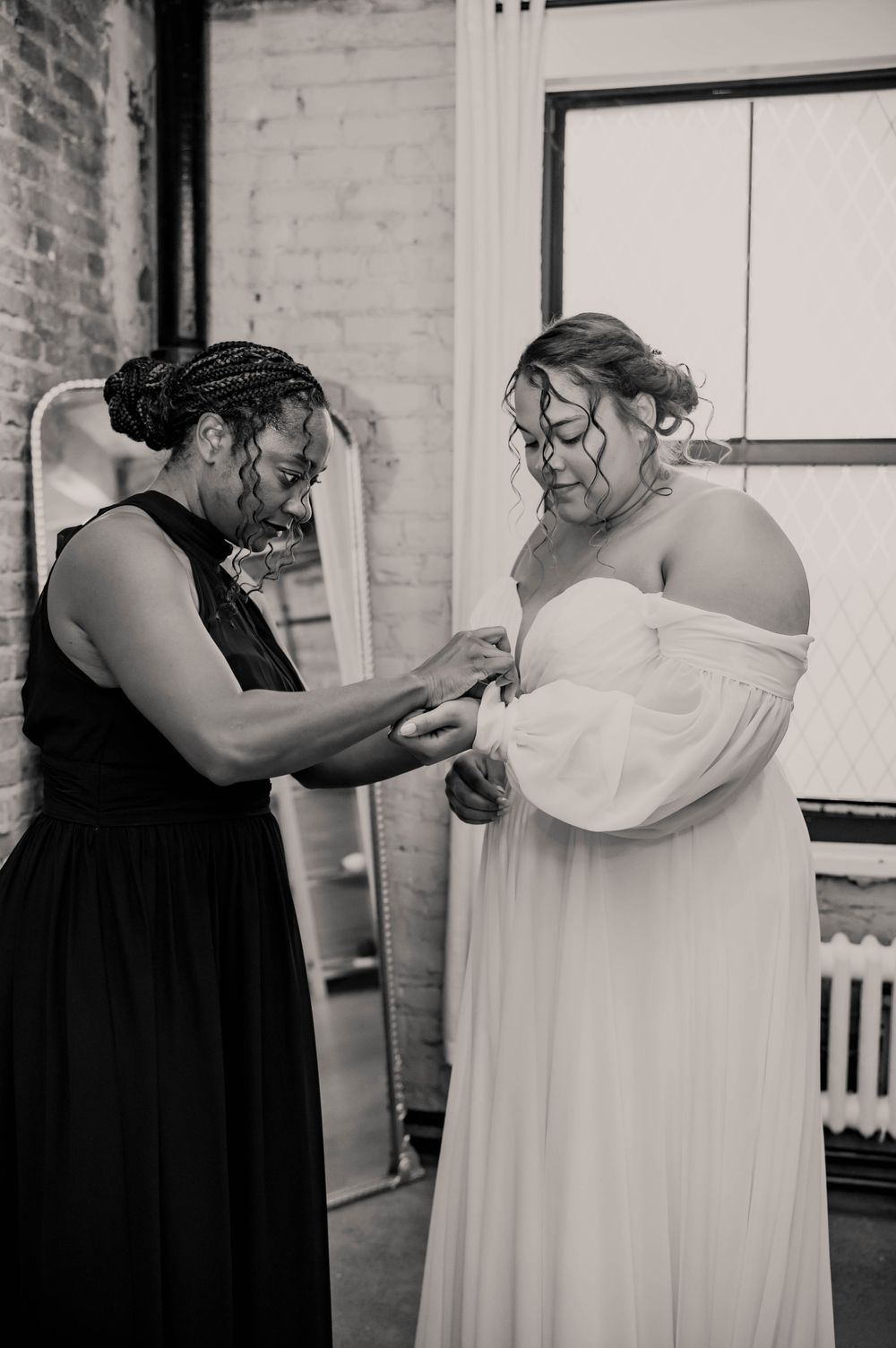Black and white photo of a woman in black dress helping a bride in an off-shoulder gown with her wedding day preparations.