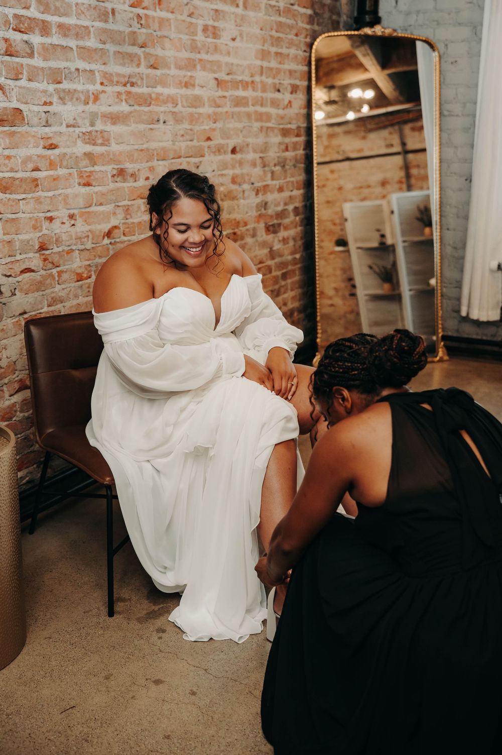 Smiling bride in off-shoulder white gown gets shoes fastened by bridesmaid in black dress near brick wall.