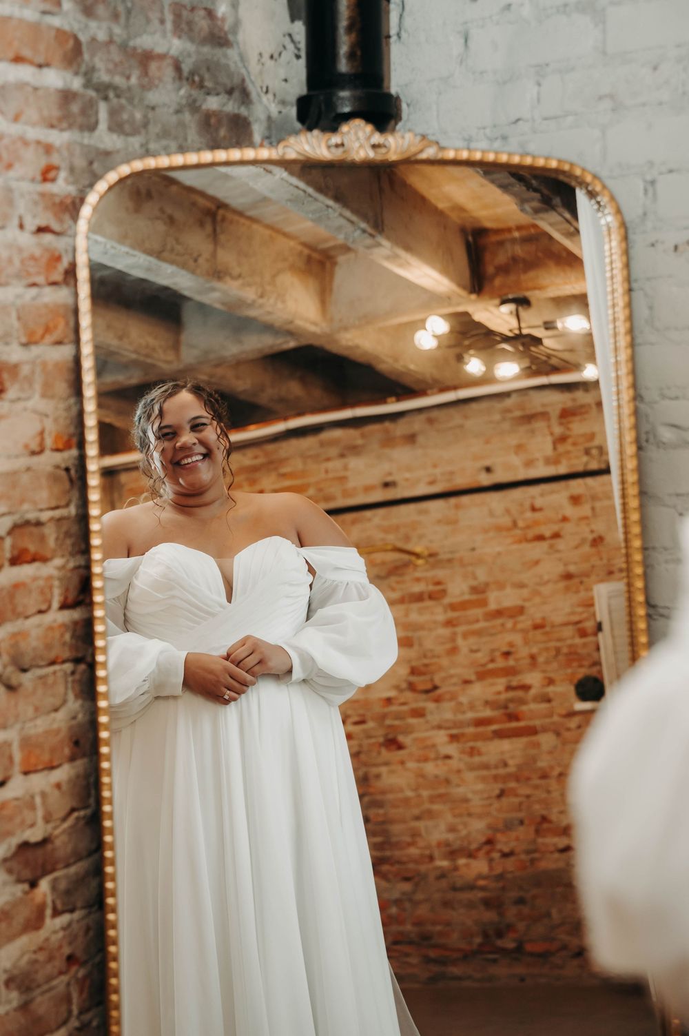 Radiant bride in off-shoulder white ballgown with puff sleeves smiling beside ornate gold floor mirror.