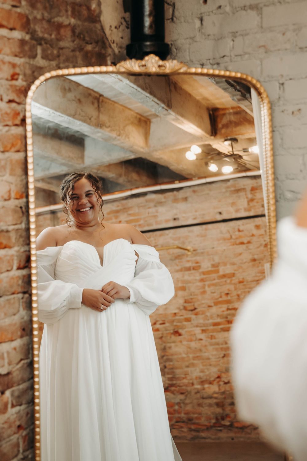 Beautiful bride in elegant white off-shoulder gown with puff sleeves posing in front of gold ornate mirror.