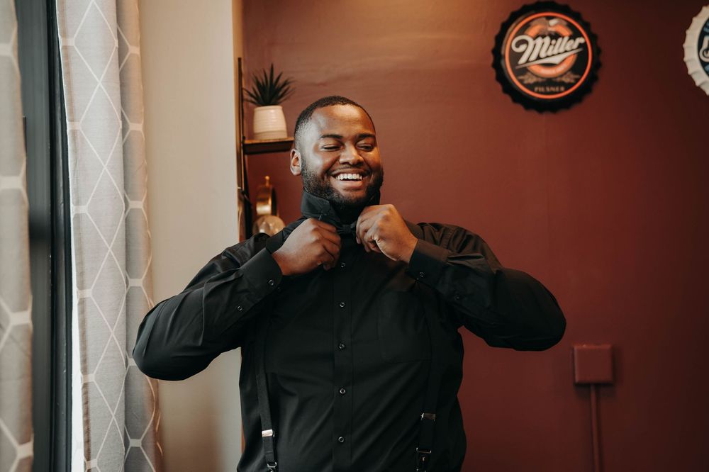 Smiling groom in black tuxedo adjusting his bow tie while getting ready for his wedding day.
