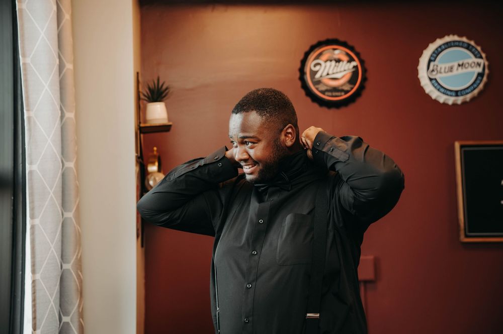 Groom in black suit laughing while adjusting his collar in a warmly decorated room before wedding.
