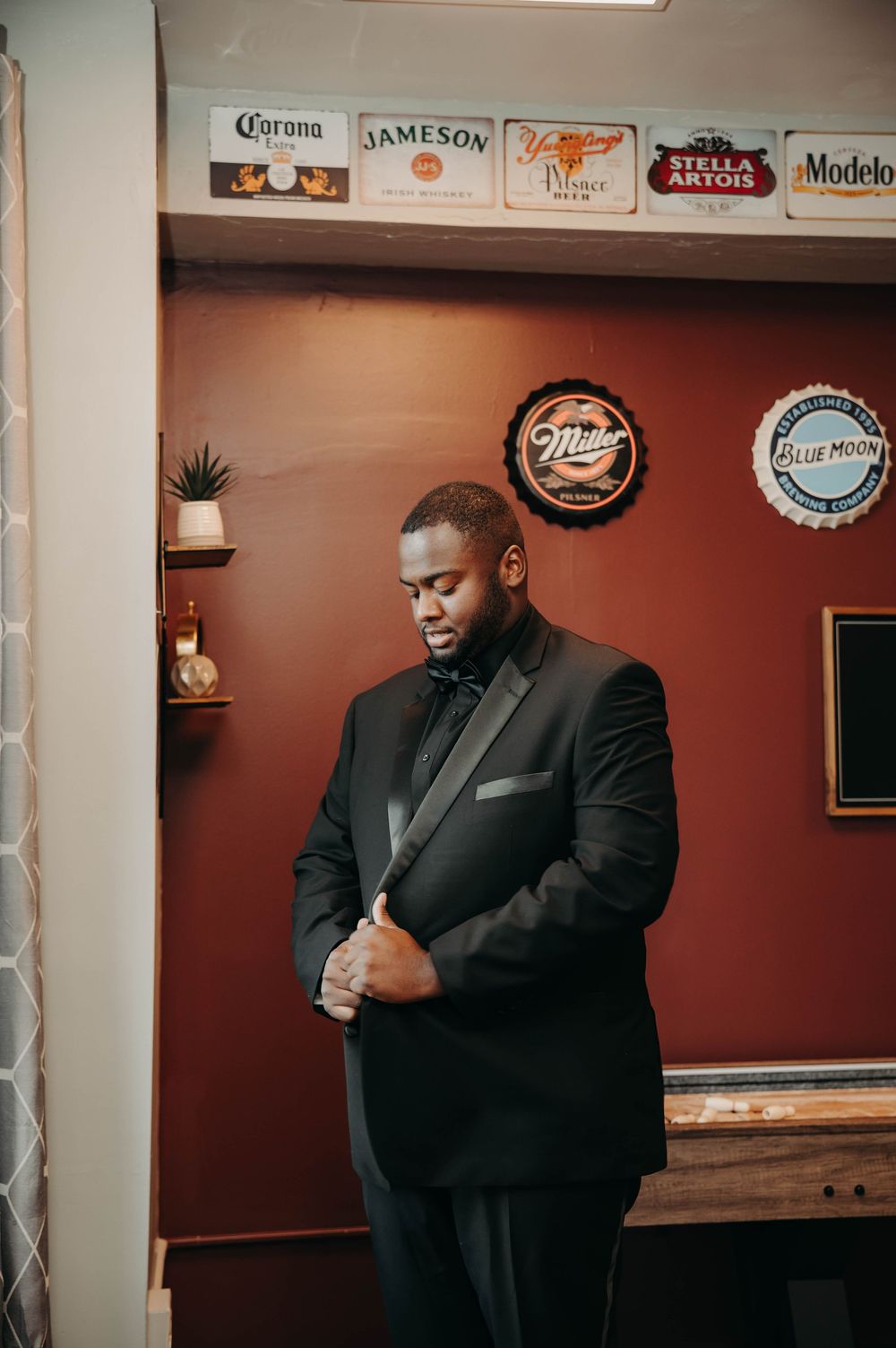 Groom in black tuxedo buttoning jacket in bar room decorated with vintage beer brand signs.