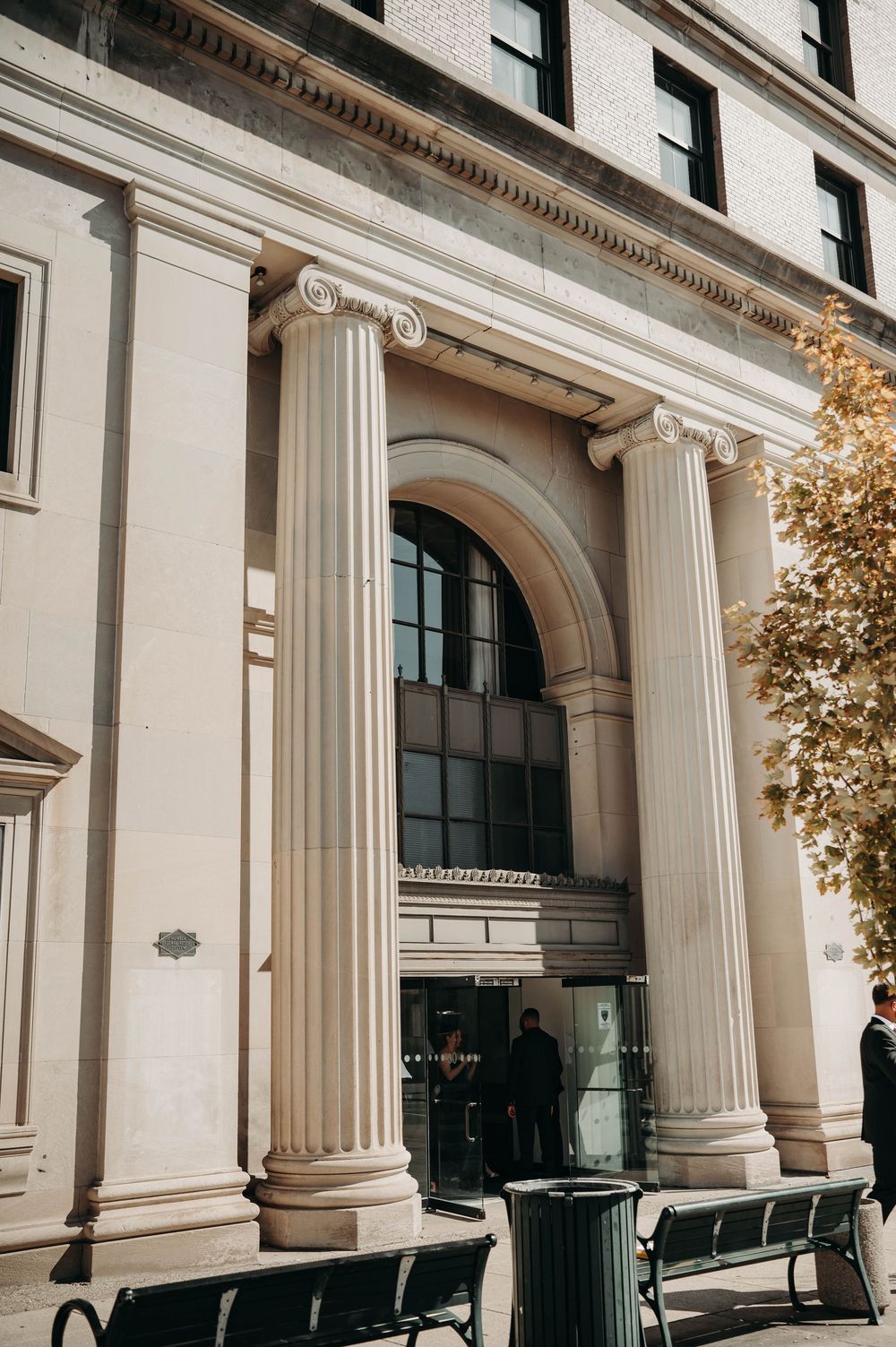 Neoclassical building entrance with tall Ionic columns, arched window, and benches in warm autumn sunlight.