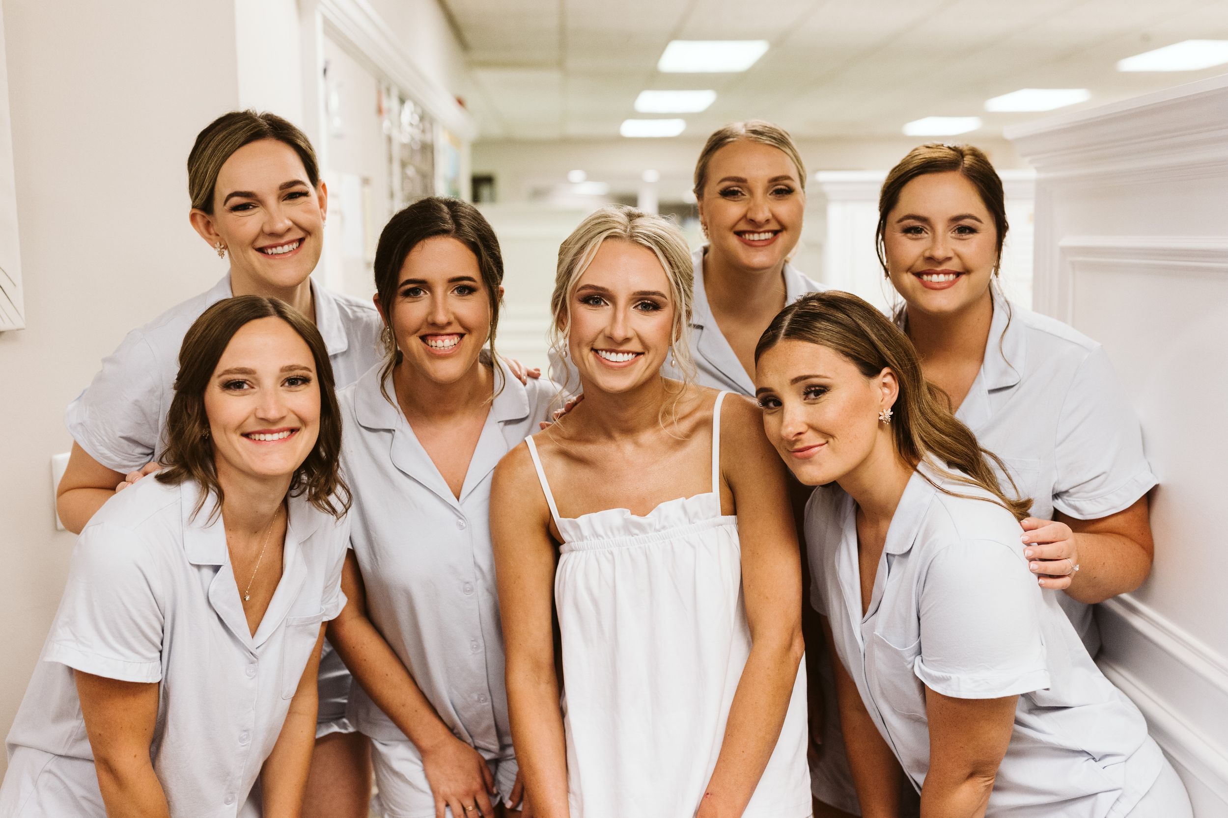 Wedding party members in white dresses pose together in a bright hallway.
