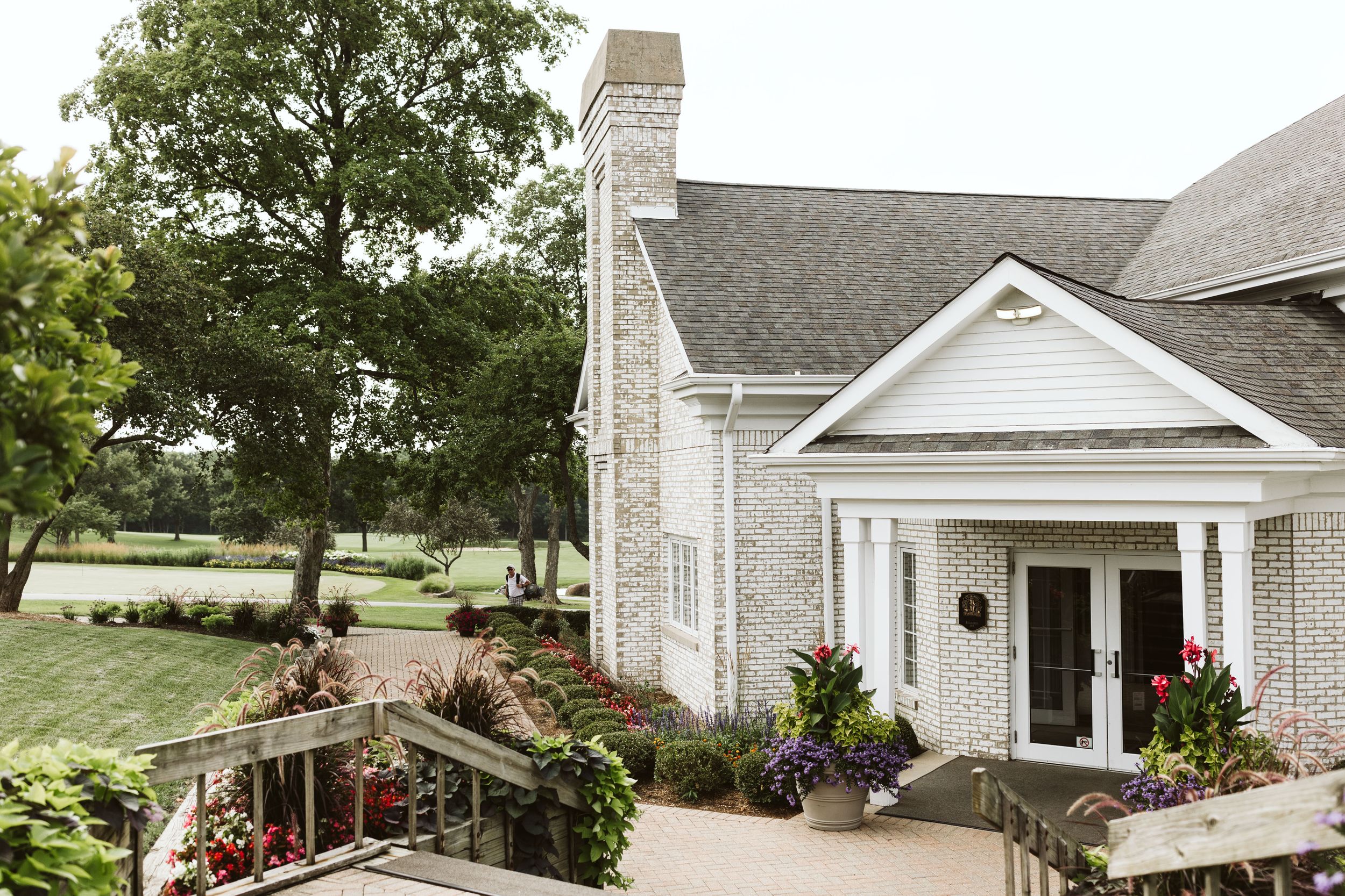 White farmhouse-style wedding venue with stone chimney, covered porch and lush landscaping on a sunny day.