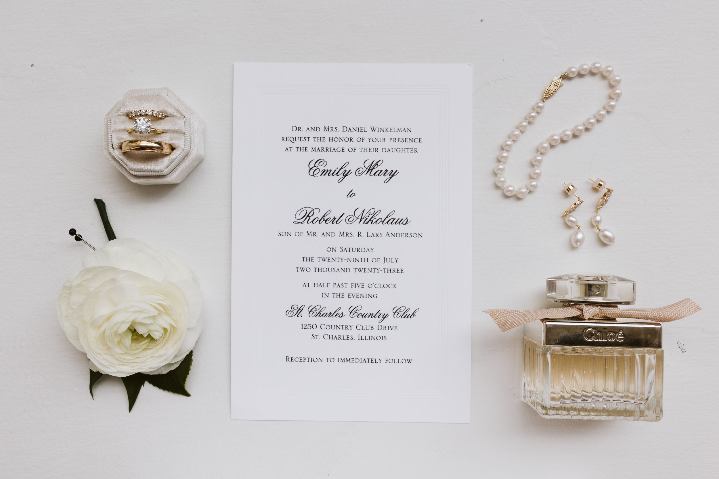 Elegant wedding details including invitations, perfume bottle, pearl jewelry and white flowers arranged on light background.
