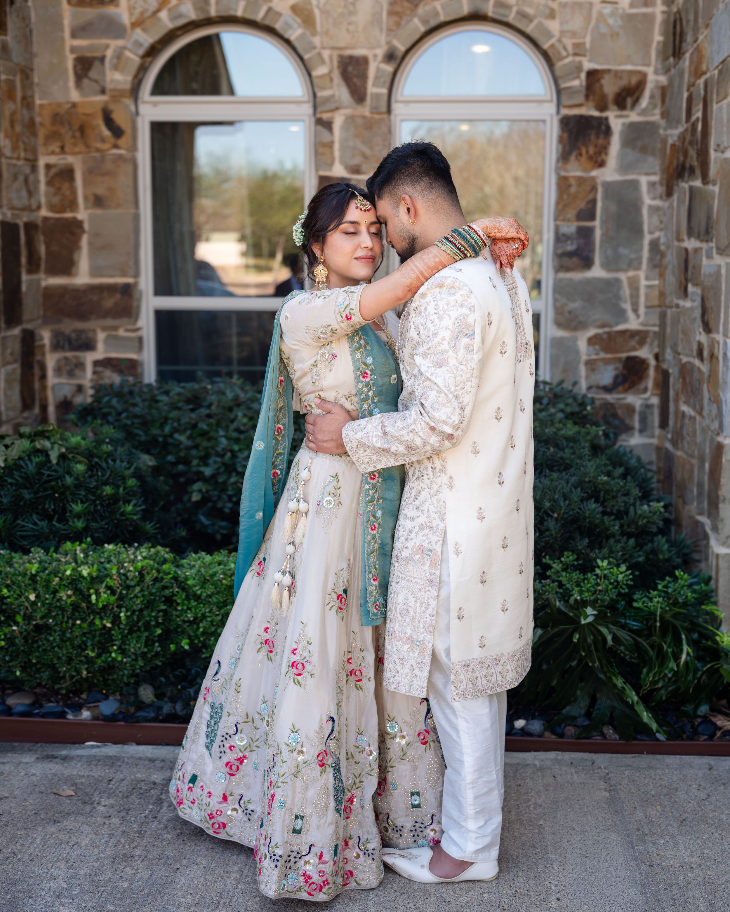 A couple in traditional South Asian wedding attire embraces while standing against a stone wall with greenery.