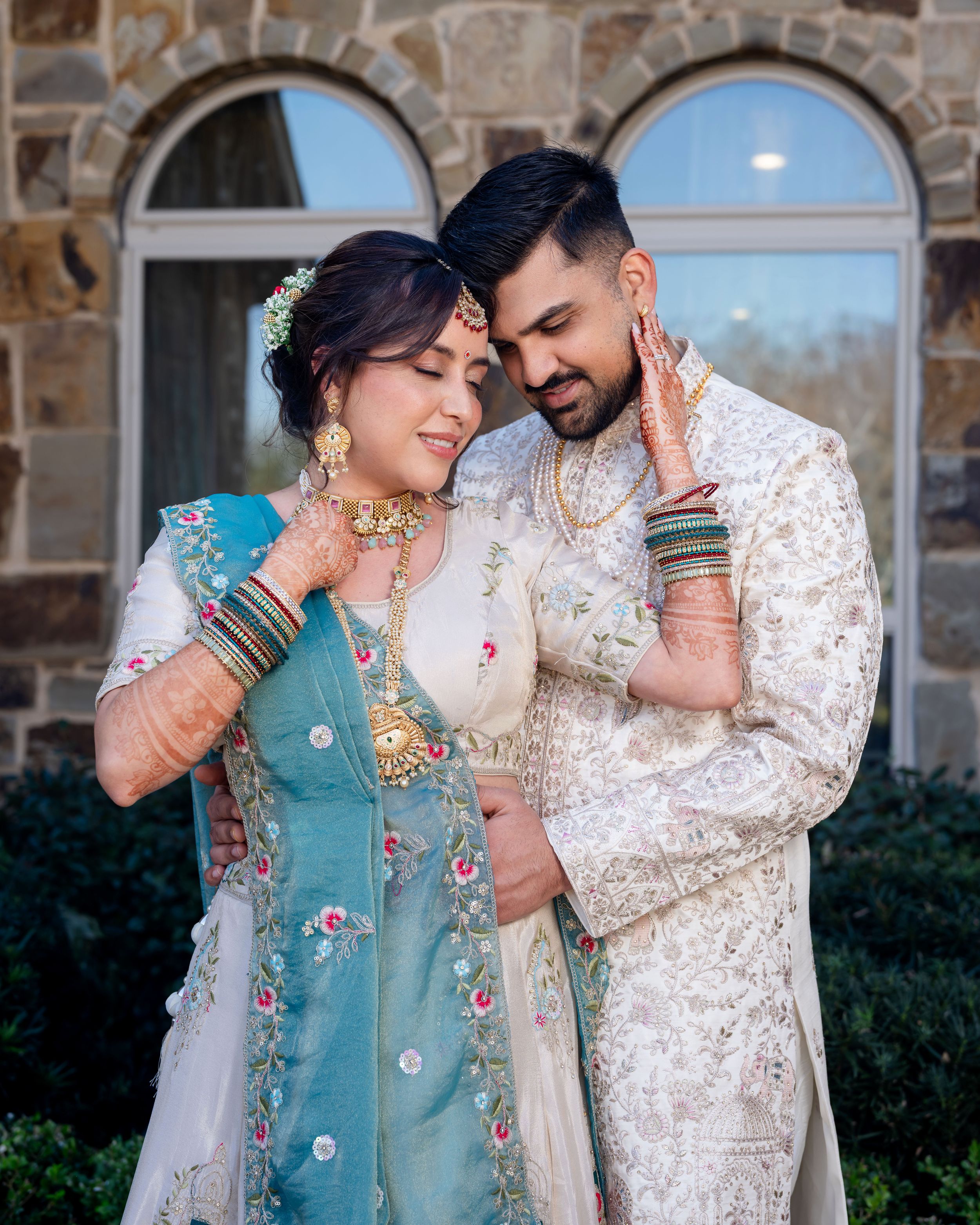 A couple in traditional South Asian attire embraces lovingly in front of a stone building during a photoshoot.