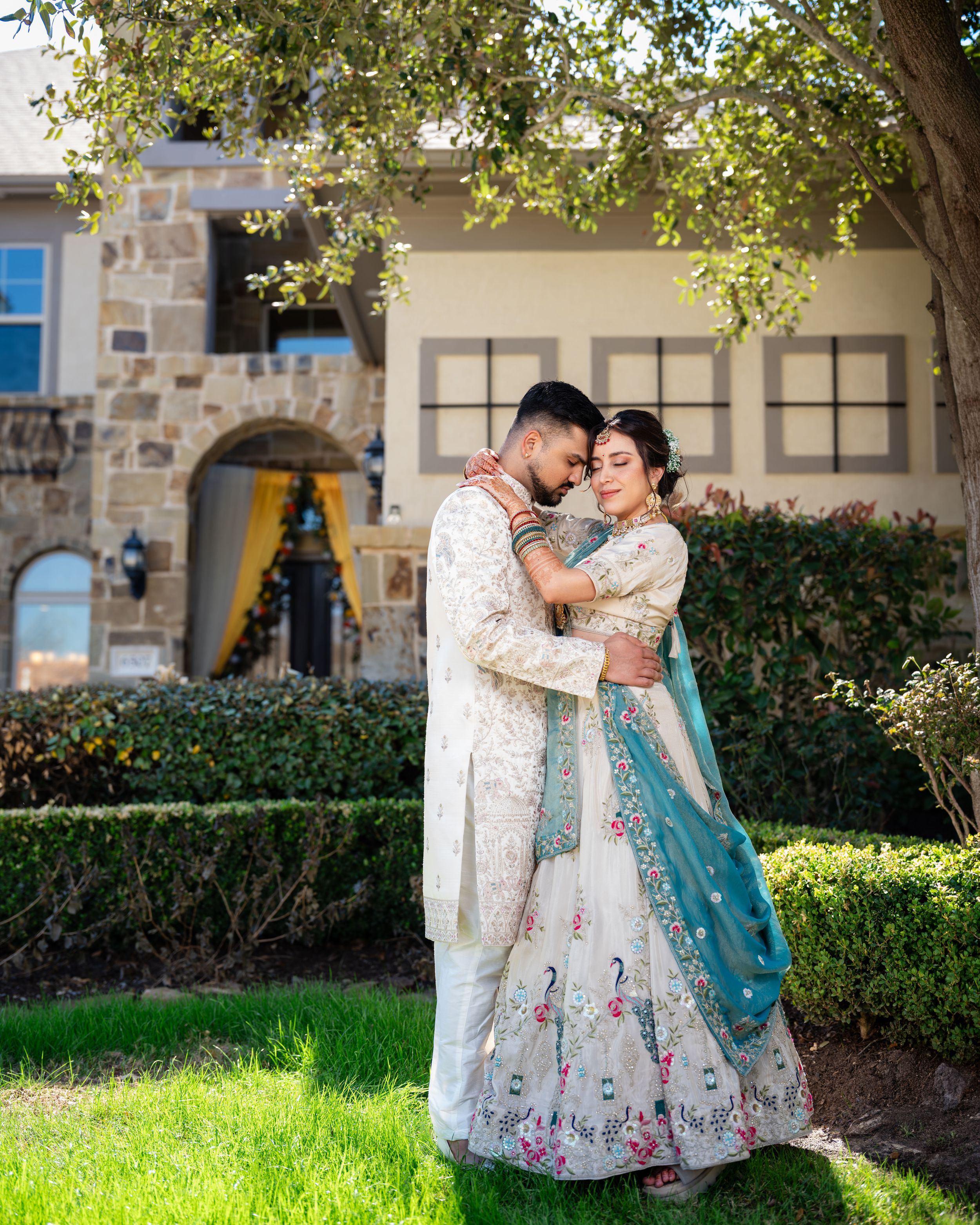 A couple in traditional South Asian attire share romantic moments in a garden setting with stone architecture.